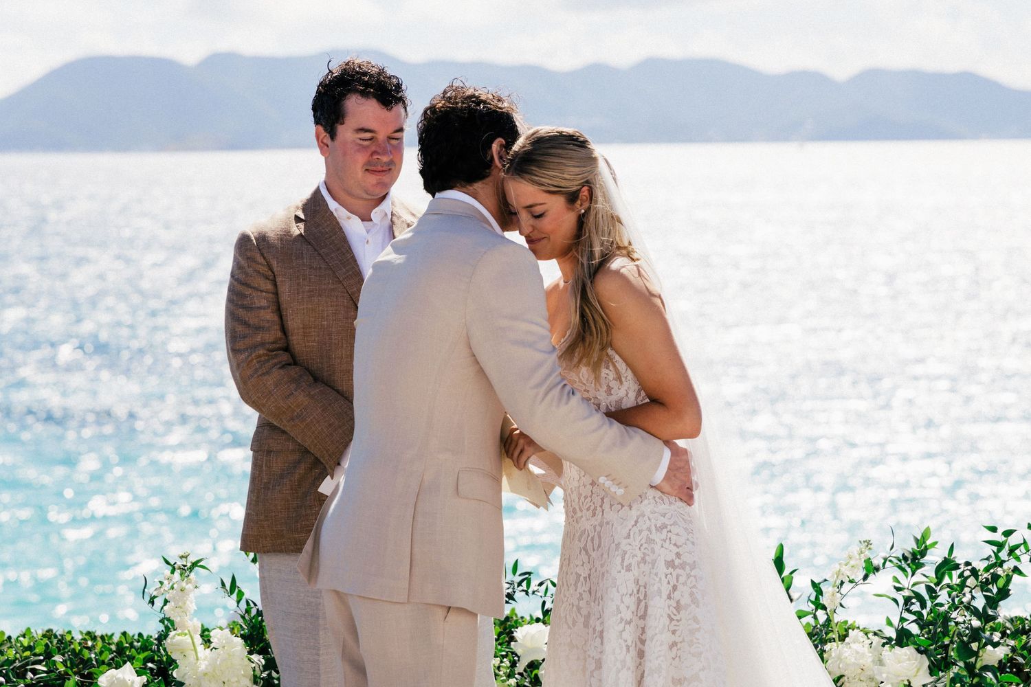 A beachside wedding ceremony with guests in light colored attire against a backdrop of turquoise ocean waters.