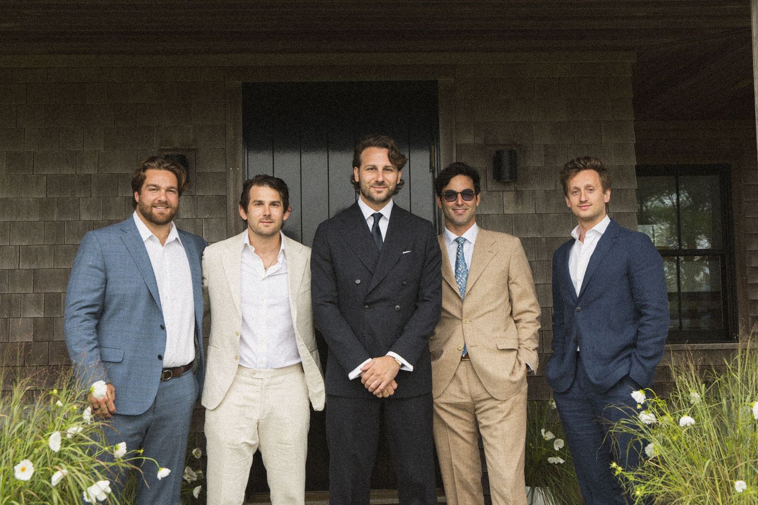 Five well-dressed men in suits posing together outdoors in front of a building surrounded by greenery.