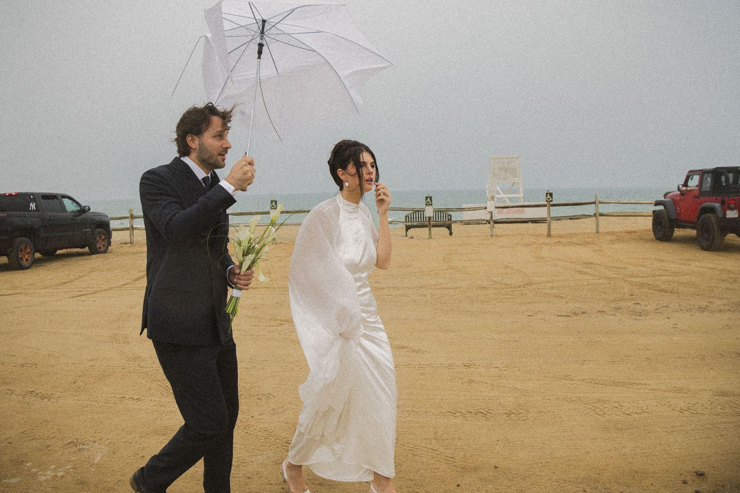 Bride and groom walking on sandy beach in rain under clear umbrella, wearing wedding attire with ocean in background.