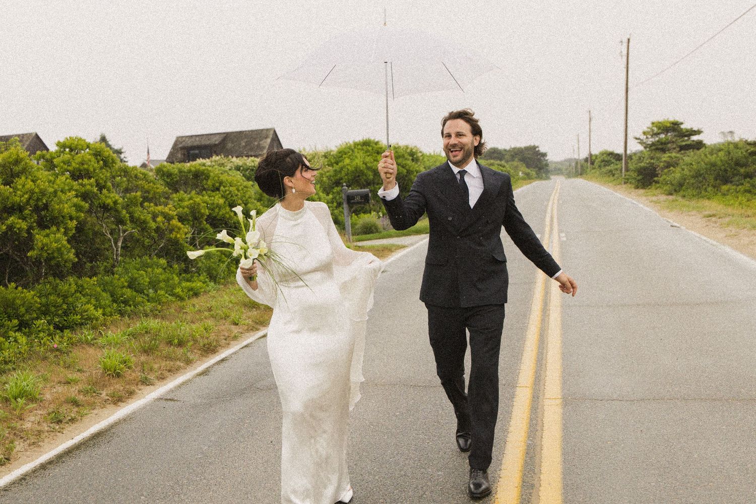 A bride and groom walk hand-in-hand on a coastal road in the rain, holding a clear umbrella on their wedding day.