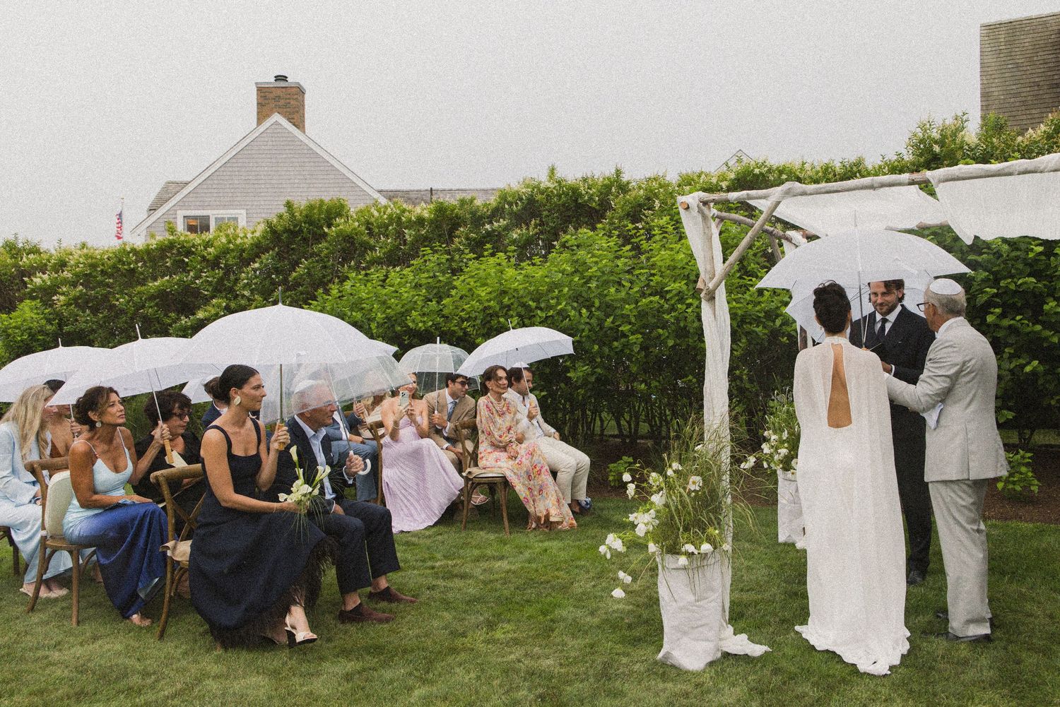 Outdoor wedding ceremony on a rainy day with guests holding white umbrellas while couple exchanges vows under a floral arch.