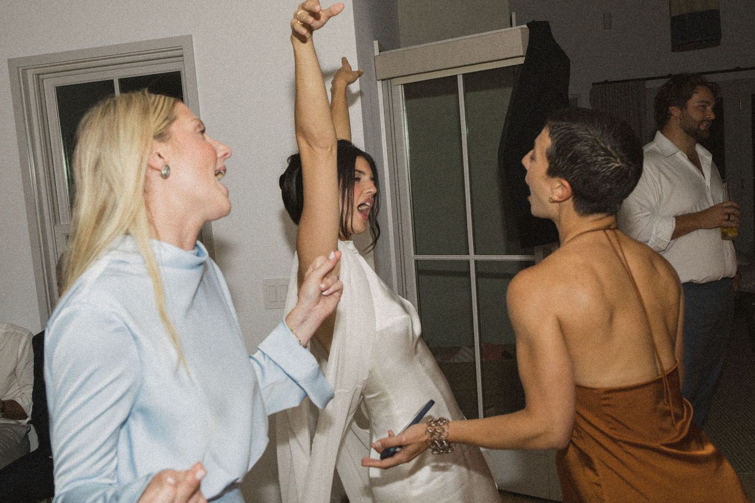 Three women dancing and celebrating together at an indoor party, wearing elegant outfits in a candid joyful moment.