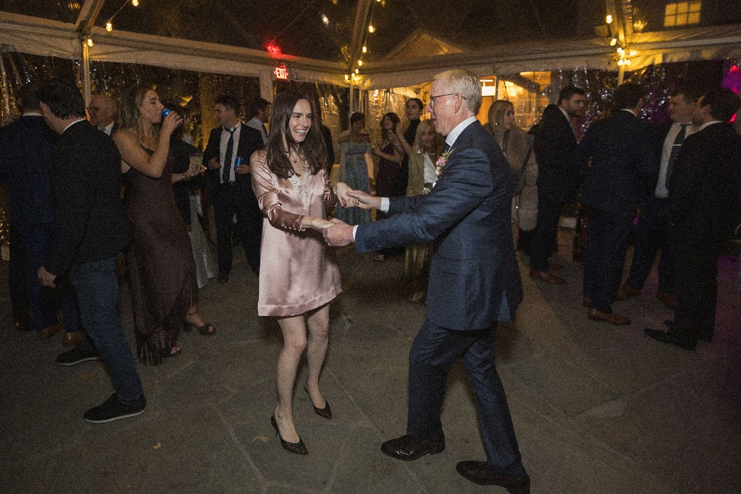 A woman in a blush dress and a gray-haired man in a navy suit dance together as wedding guests mingle under string lights.