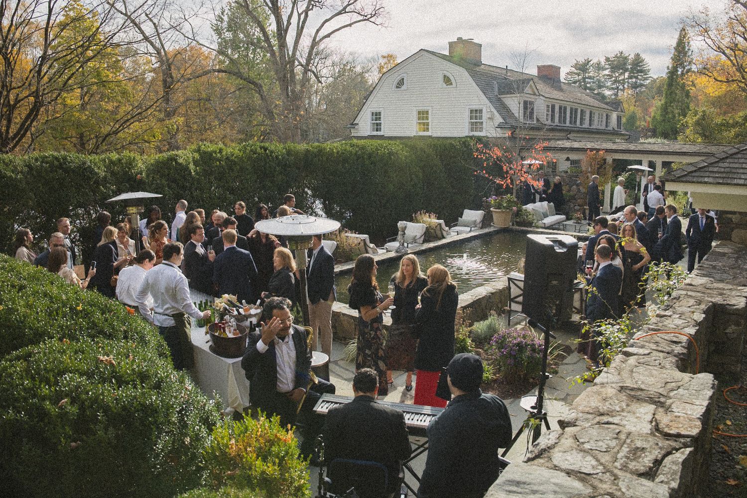 Wedding guests mingle during an outdoor cocktail reception on a stone terrace with autumn trees and a white farmhouse behind.
