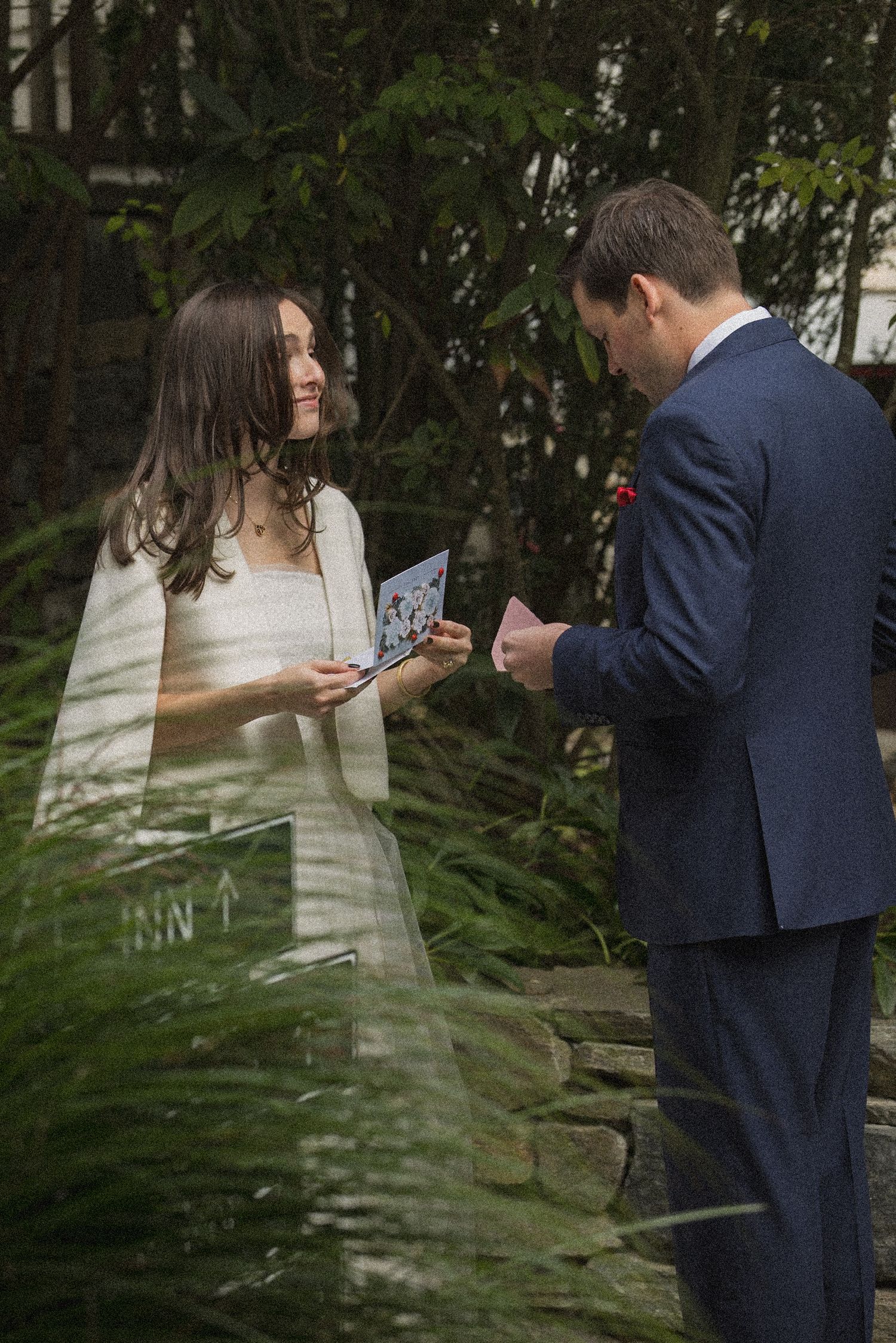 Bride in a white dress and cape and groom in a navy suit read handwritten vows to each other in a lush garden during an outdoor wedding.