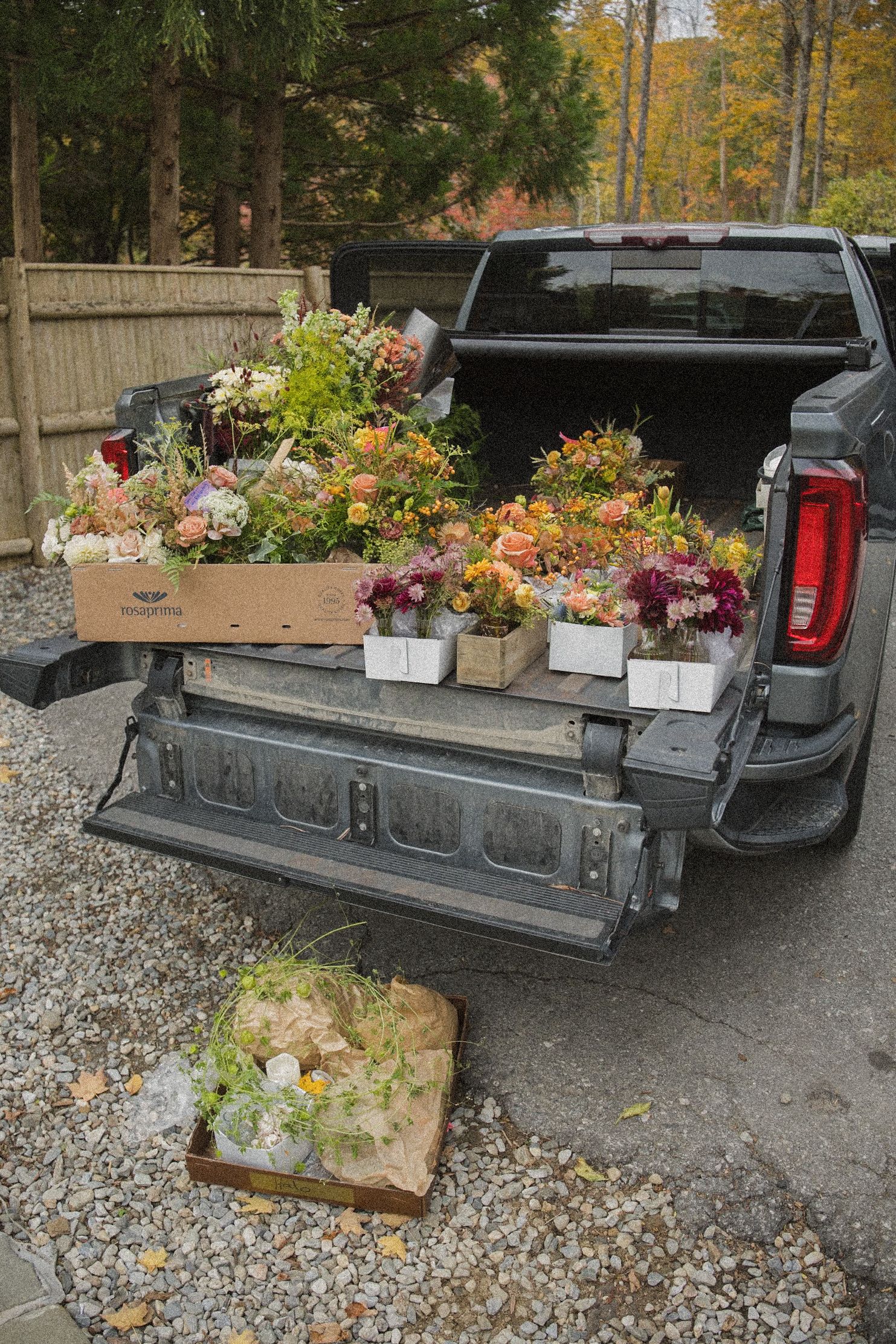 Open pickup truck bed filled with boxes of fresh orange, pink and white flowers parked on a gravel driveway surrounded by autumn trees.