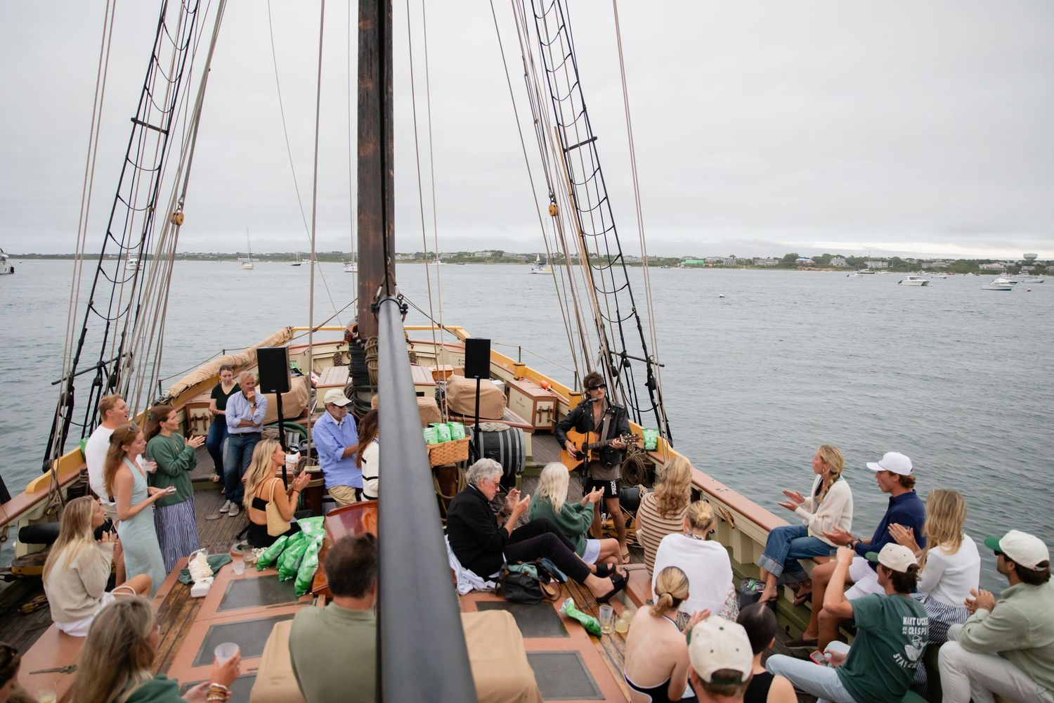 Guests gather on the deck of a tall ship sailing on calm waters under an overcast sky.