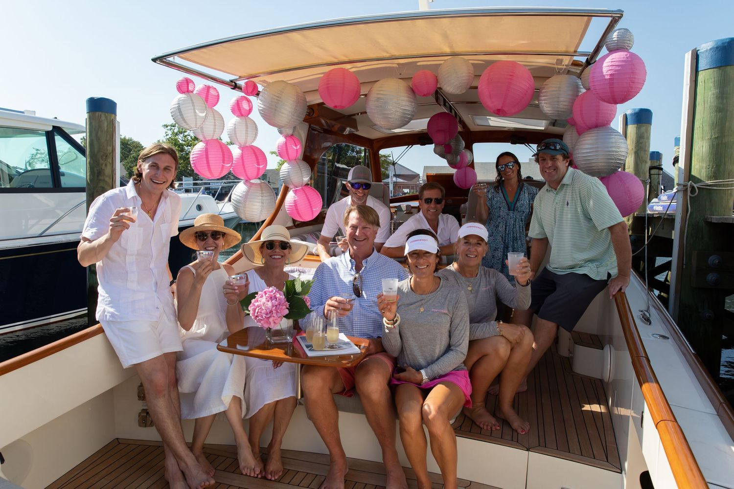 Group of friends celebrating on a decorated yacht with pink and white paper lanterns, enjoying drinks on a sunny day.