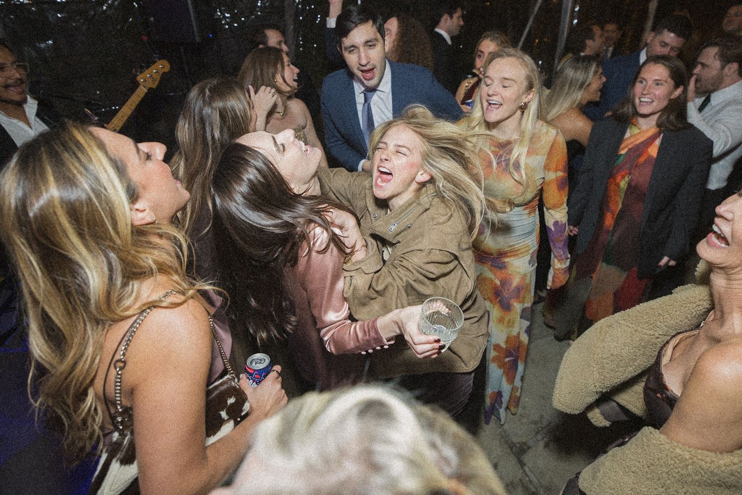 Guests in festive attire laugh and embrace while dancing in a lively crowd at an evening wedding reception.
