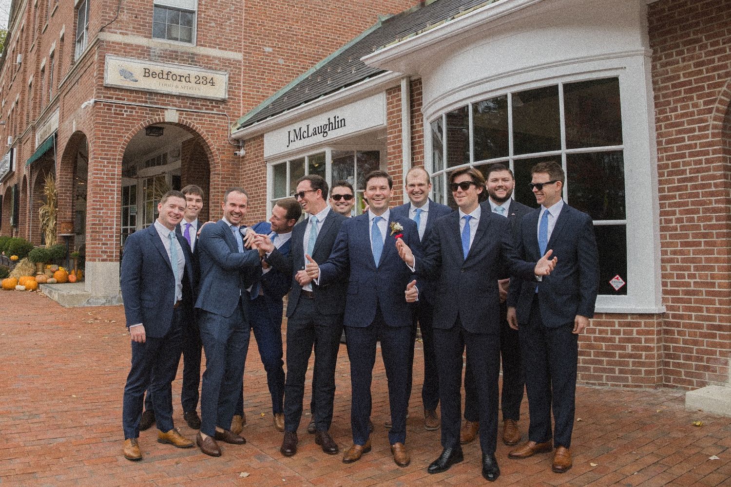 Group of groomsmen in navy suits laughing together on a brick sidewalk in front of a brick storefront.