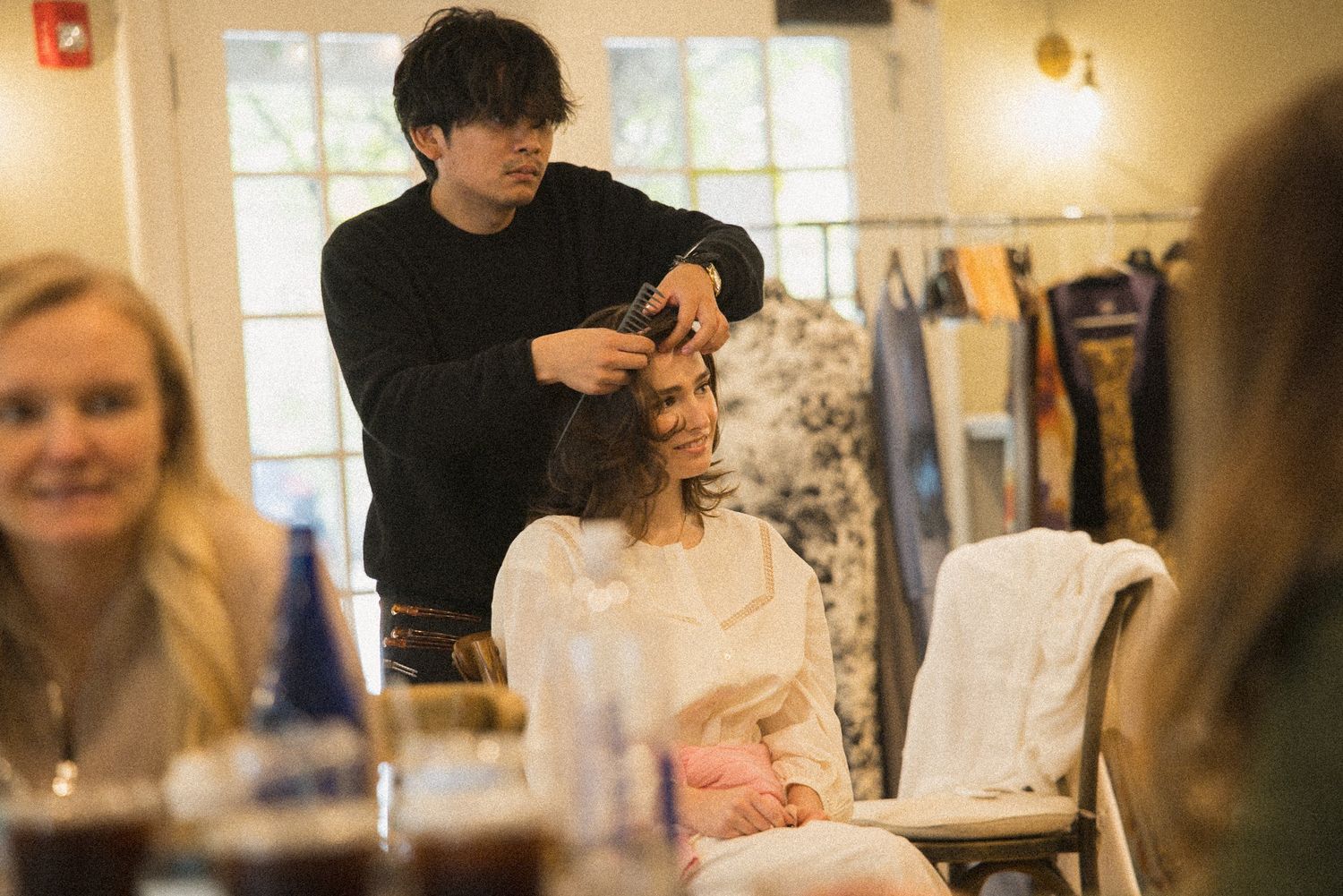 Hairstylist in a black sweater styles a seated woman's hair in a bright boutique with clothing racks while a smiling woman looks on.