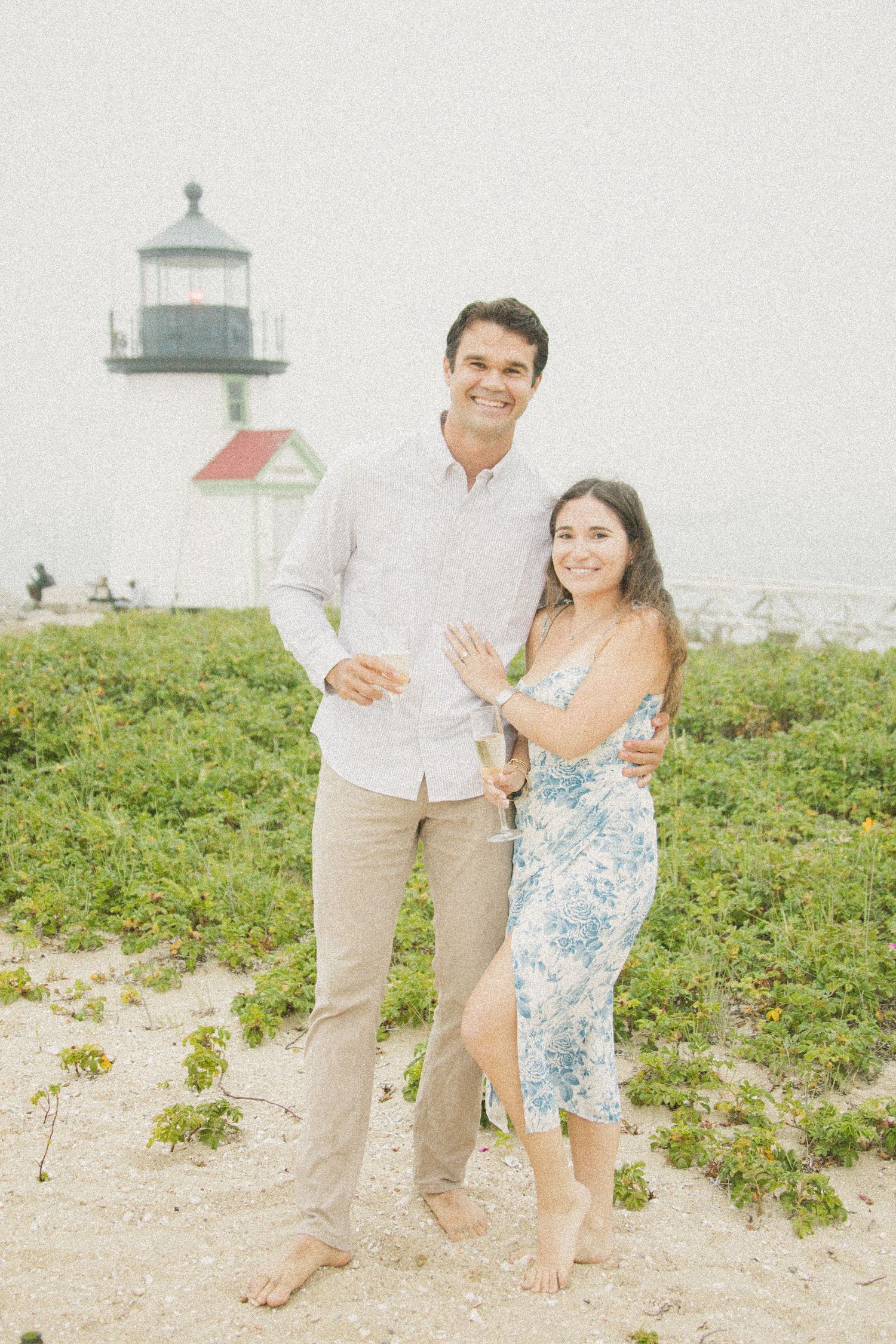 Couple smiling together on sandy beach with historic lighthouse backdrop, woman wearing floral dress, man in white shirt.