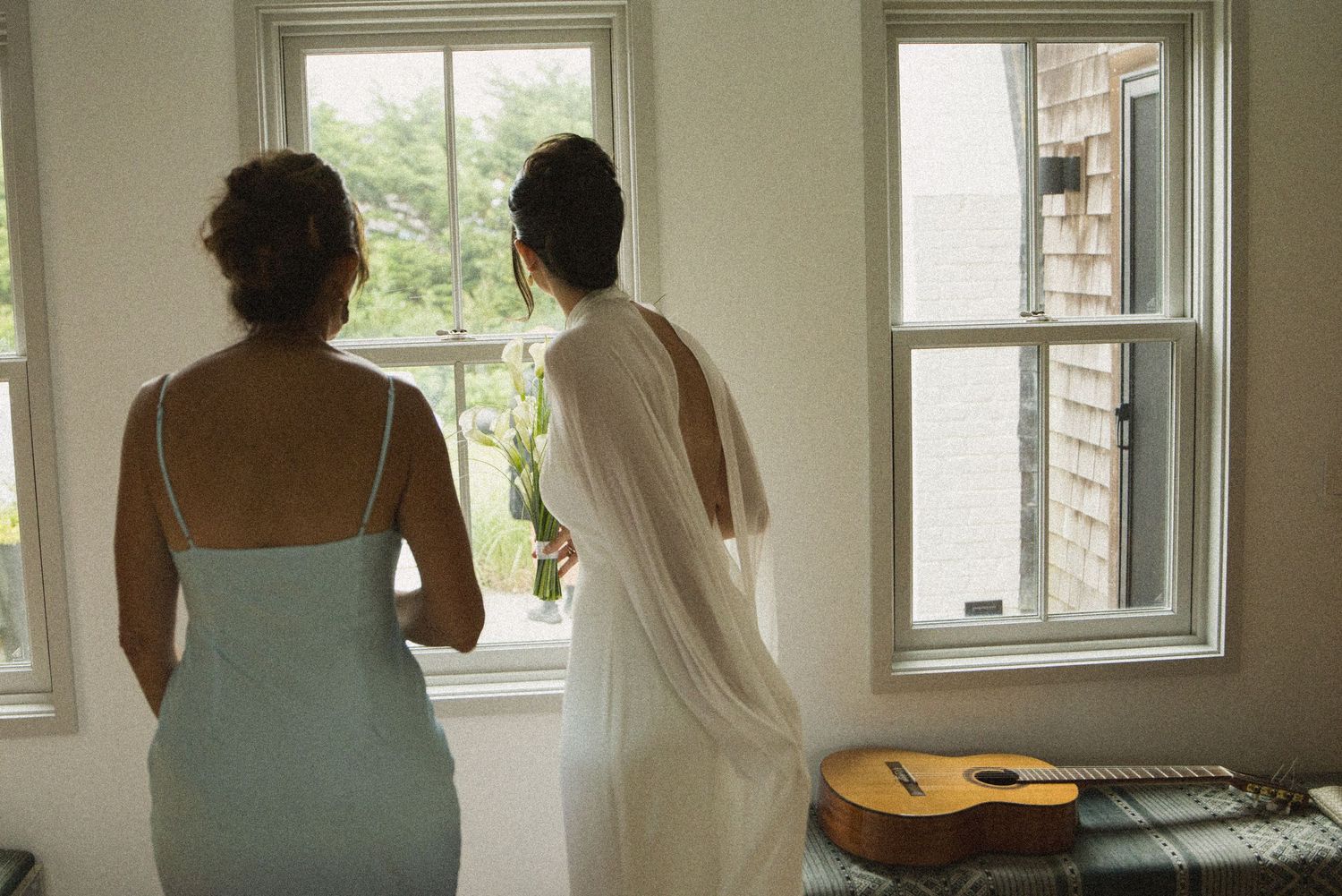 Two women stand looking out windows in a bright room with a acoustic guitar leaning against the wall.