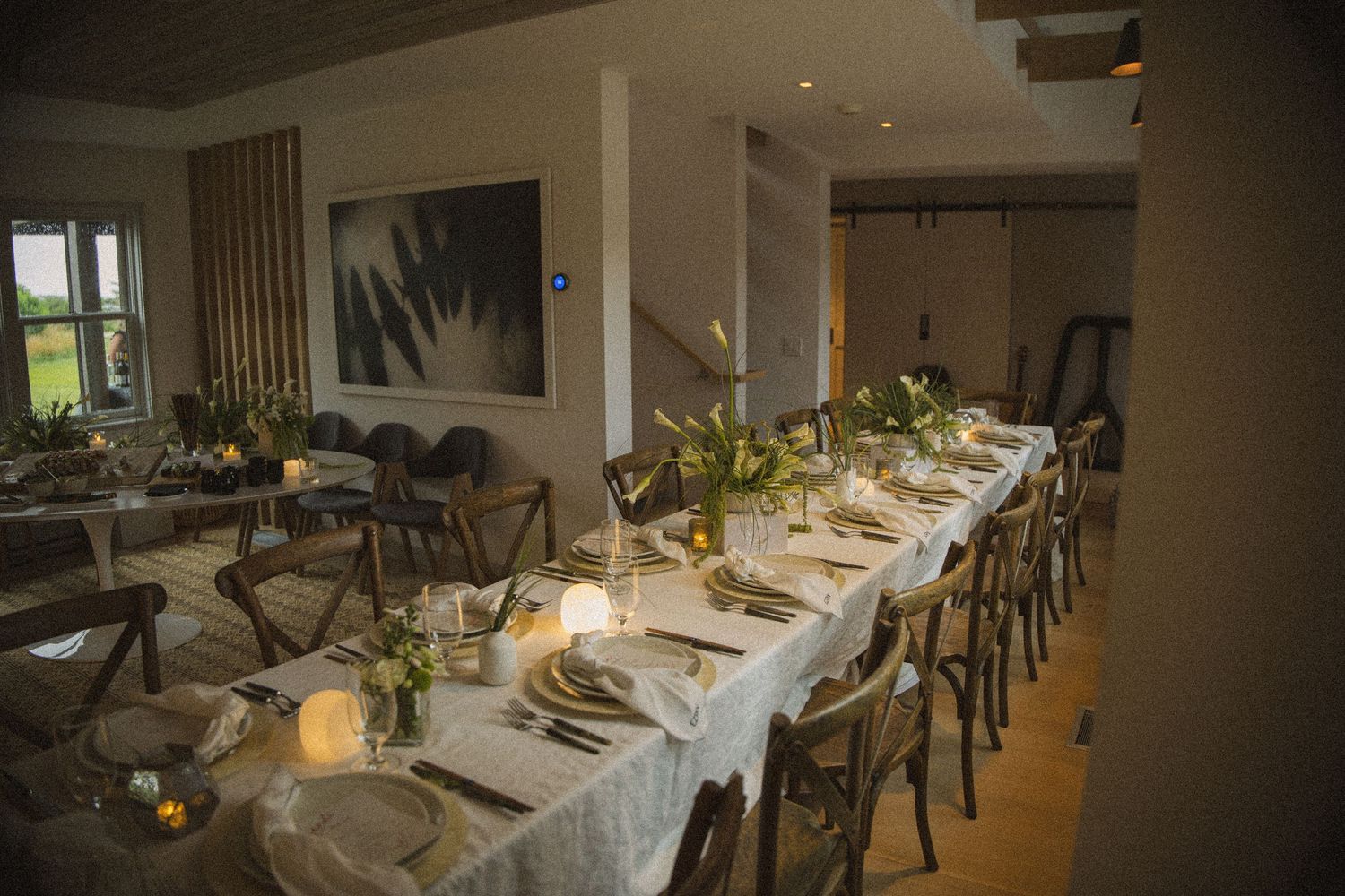 Elegant long dining table set for a formal event with candles, floral centerpieces, and rustic wooden chairs in a modern venue.