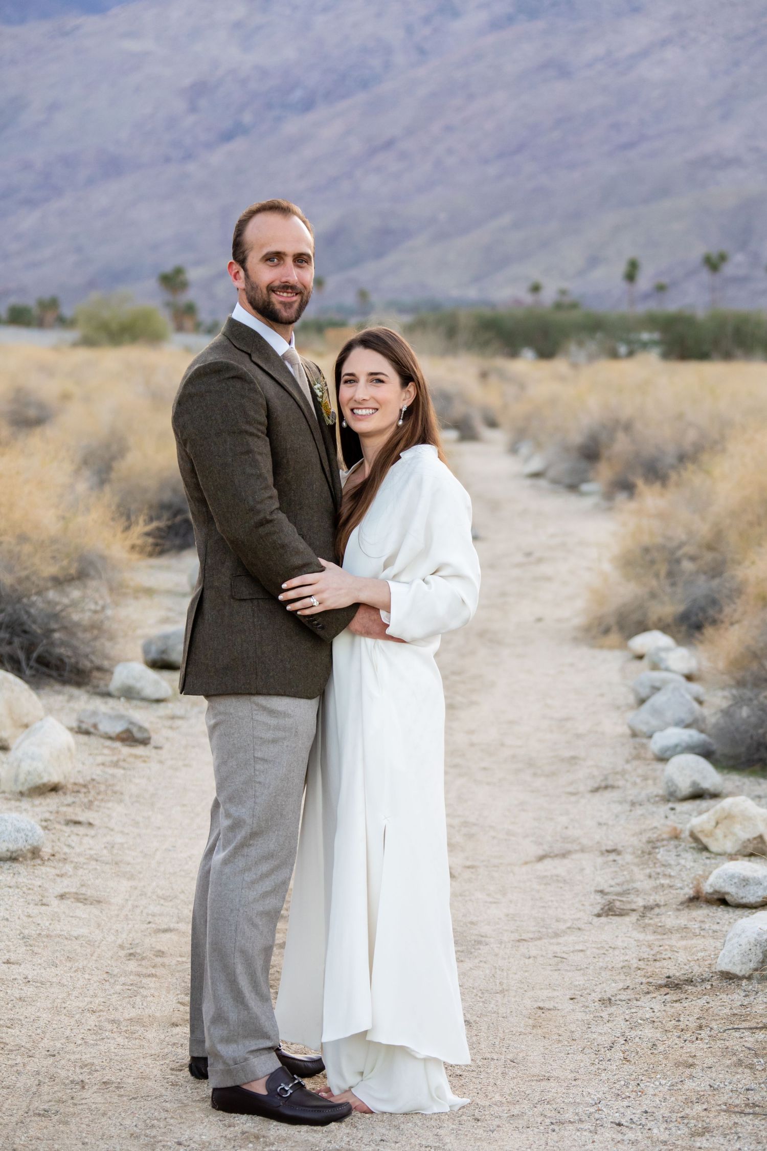 A couple embraces on a desert trail with mountains and brush in the background.