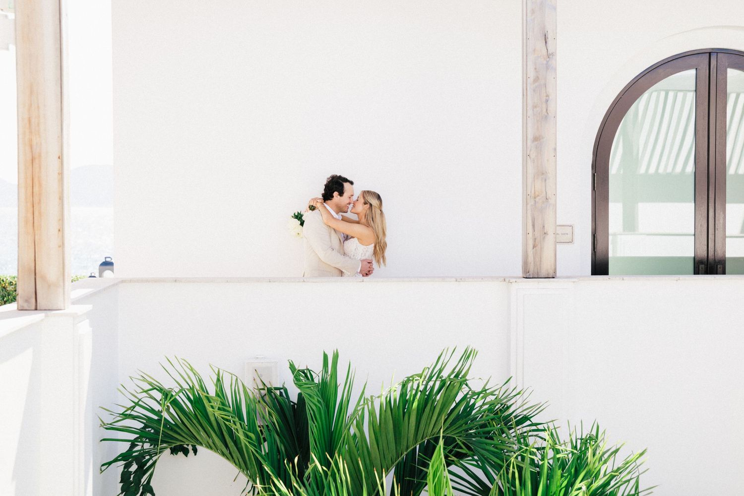 A romantic wedding portrait on a white balcony with arched windows and tropical greenery.