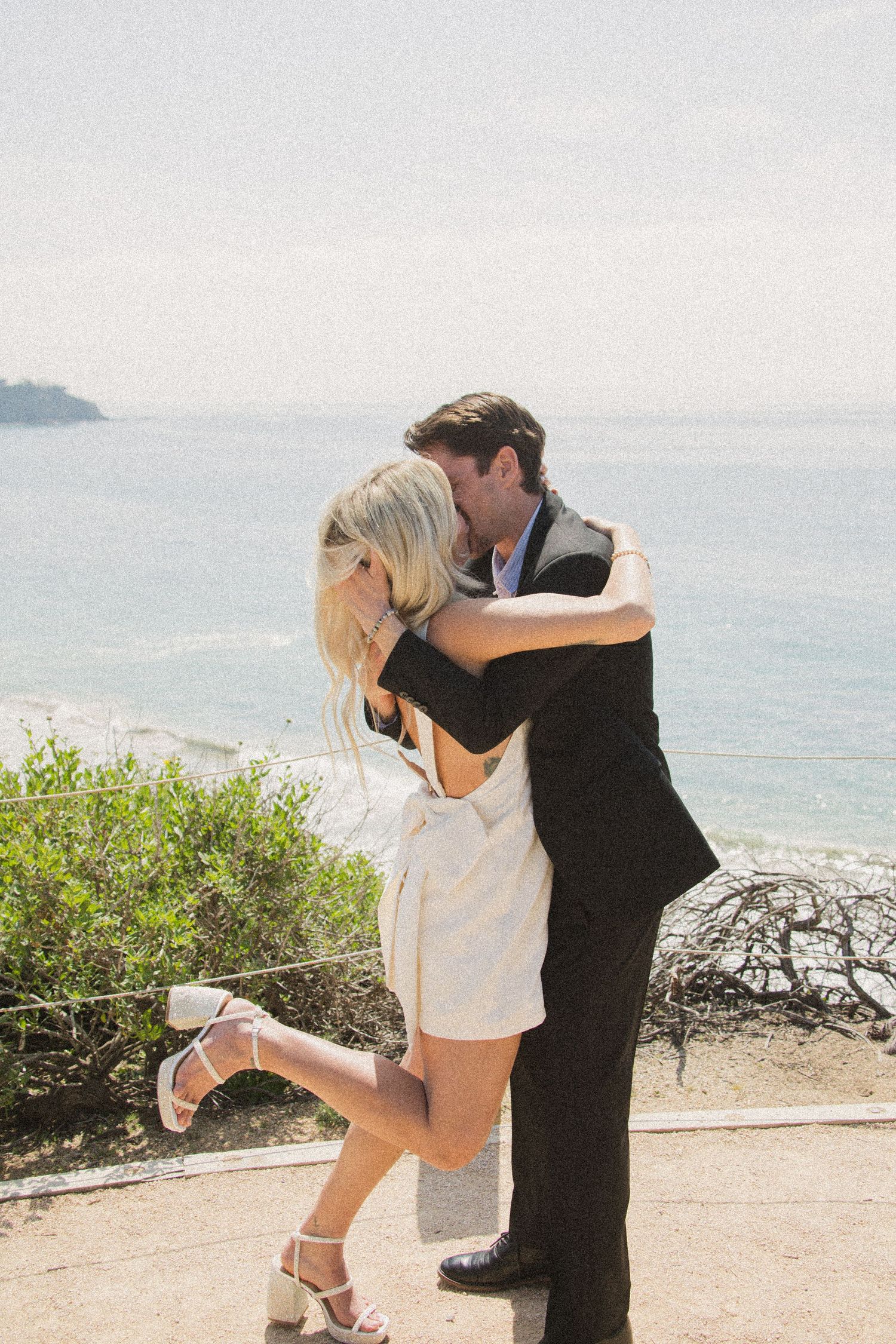 A romantic seaside photoshoot showing a couple embracing on a coastal cliff with ocean views.