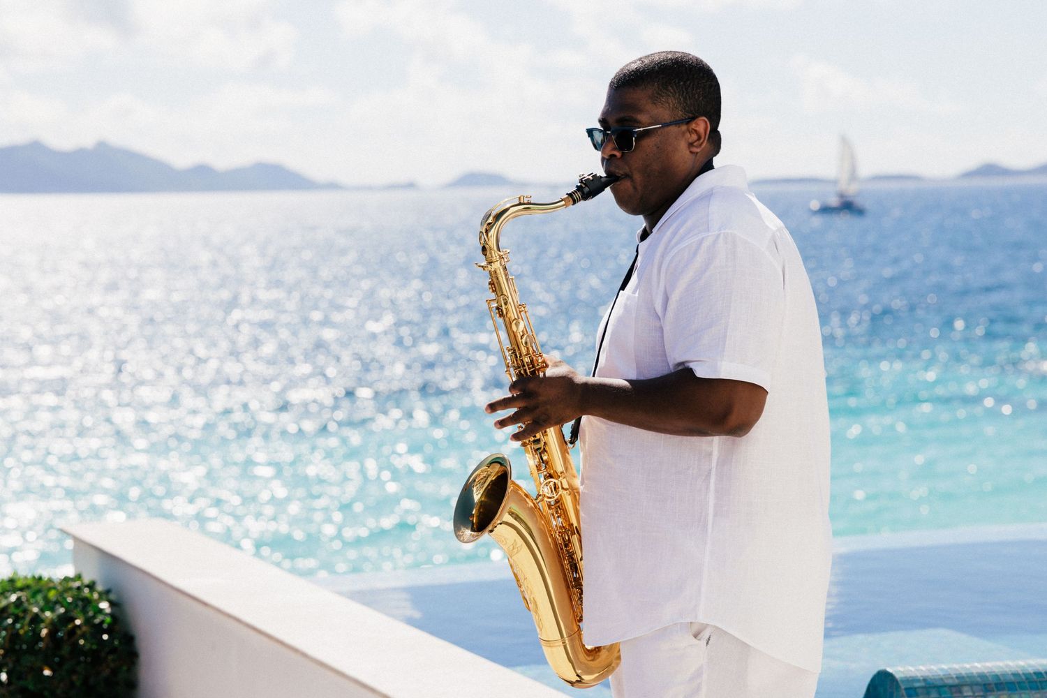 Saxophonist in white clothing performs on a seaside terrace with sparkling blue ocean and mountains in background.