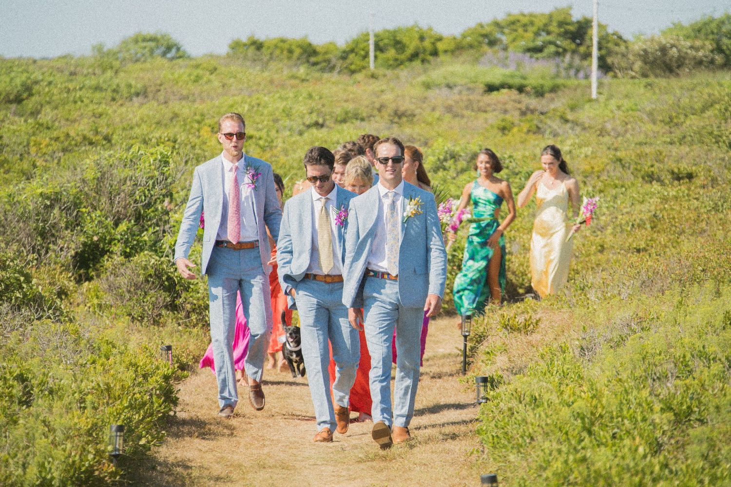 Wedding guests in colorful attire walking along green coastal path.
