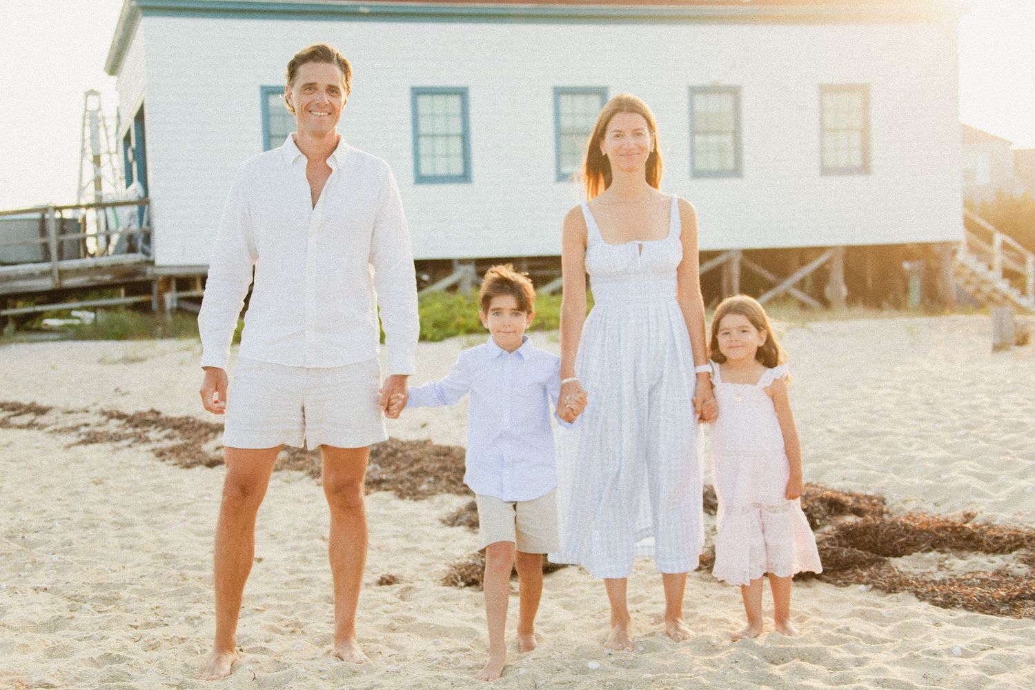 Family of four in white outfits posing on a sunny beach near a coastal building.
