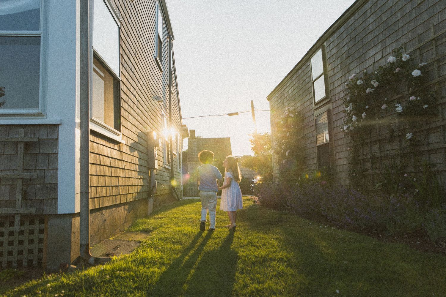 Two children walk hand-in-hand between rustic shingled houses toward a glowing sunset on a grassy path.
