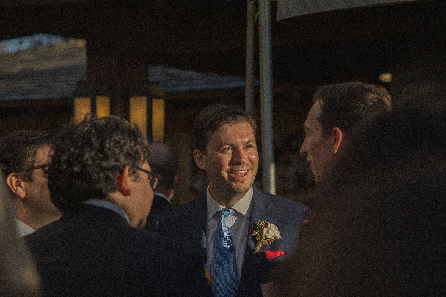 Smiling man in a navy suit with a floral boutonniere chats with other guests at an outdoor evening wedding reception.