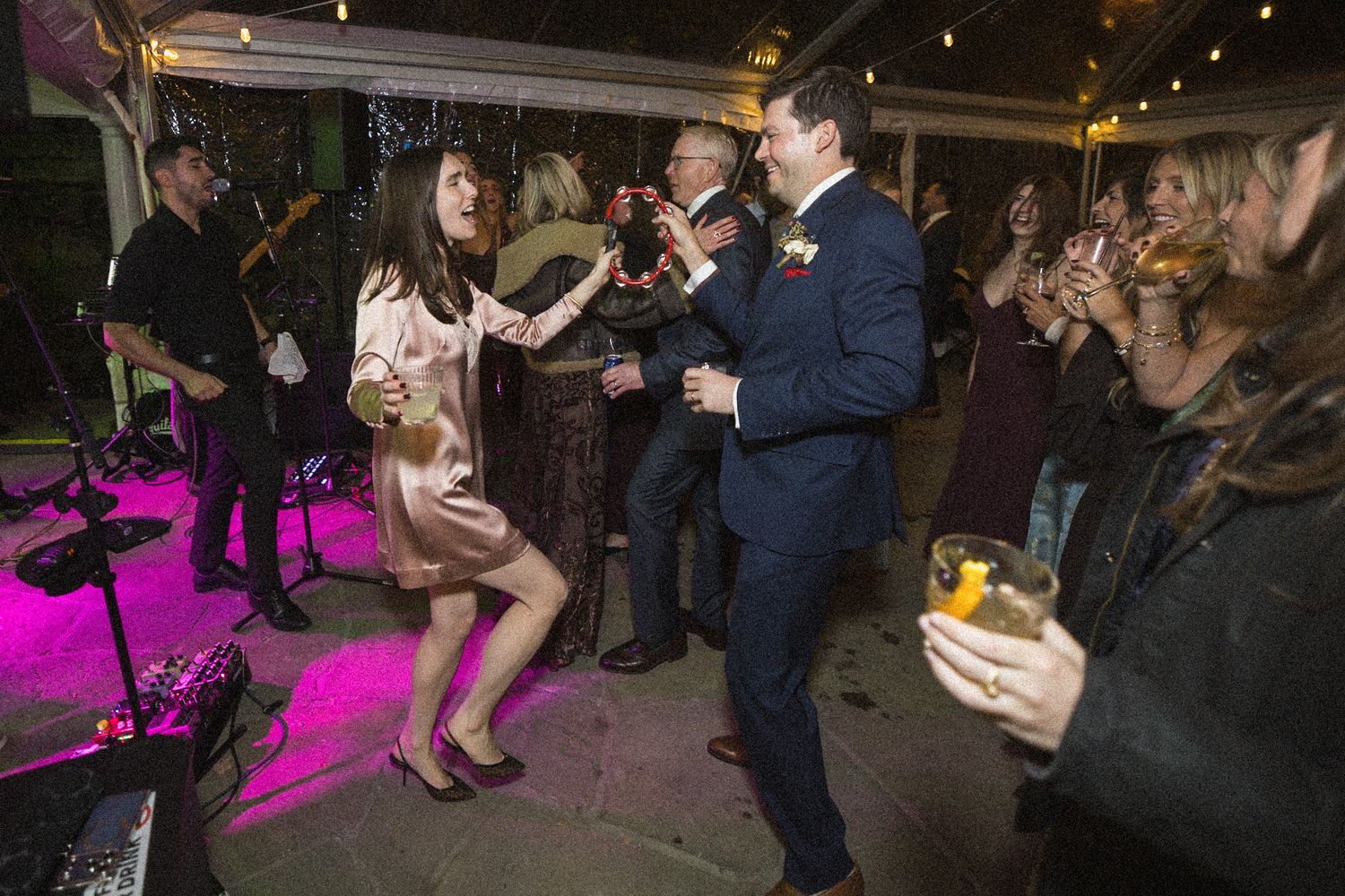 A woman in a blush dress dances with a man in a navy suit as wedding guests cheer with drinks near a live band.