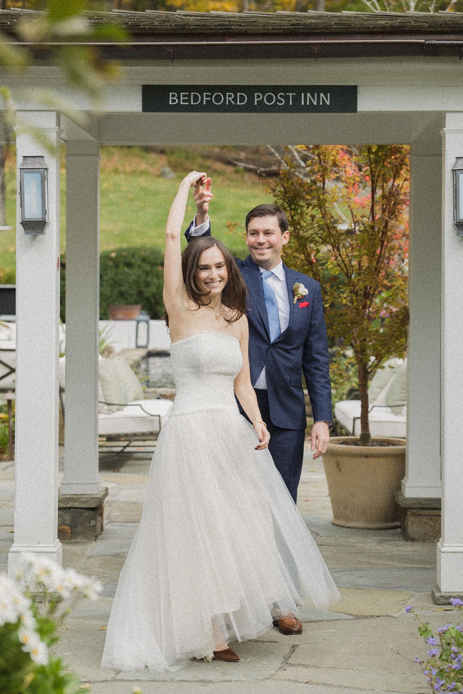 Bride in a flowing tulle gown twirls with her smiling groom beneath the Bedford Post Inn entrance on a sunny autumn wedding day.