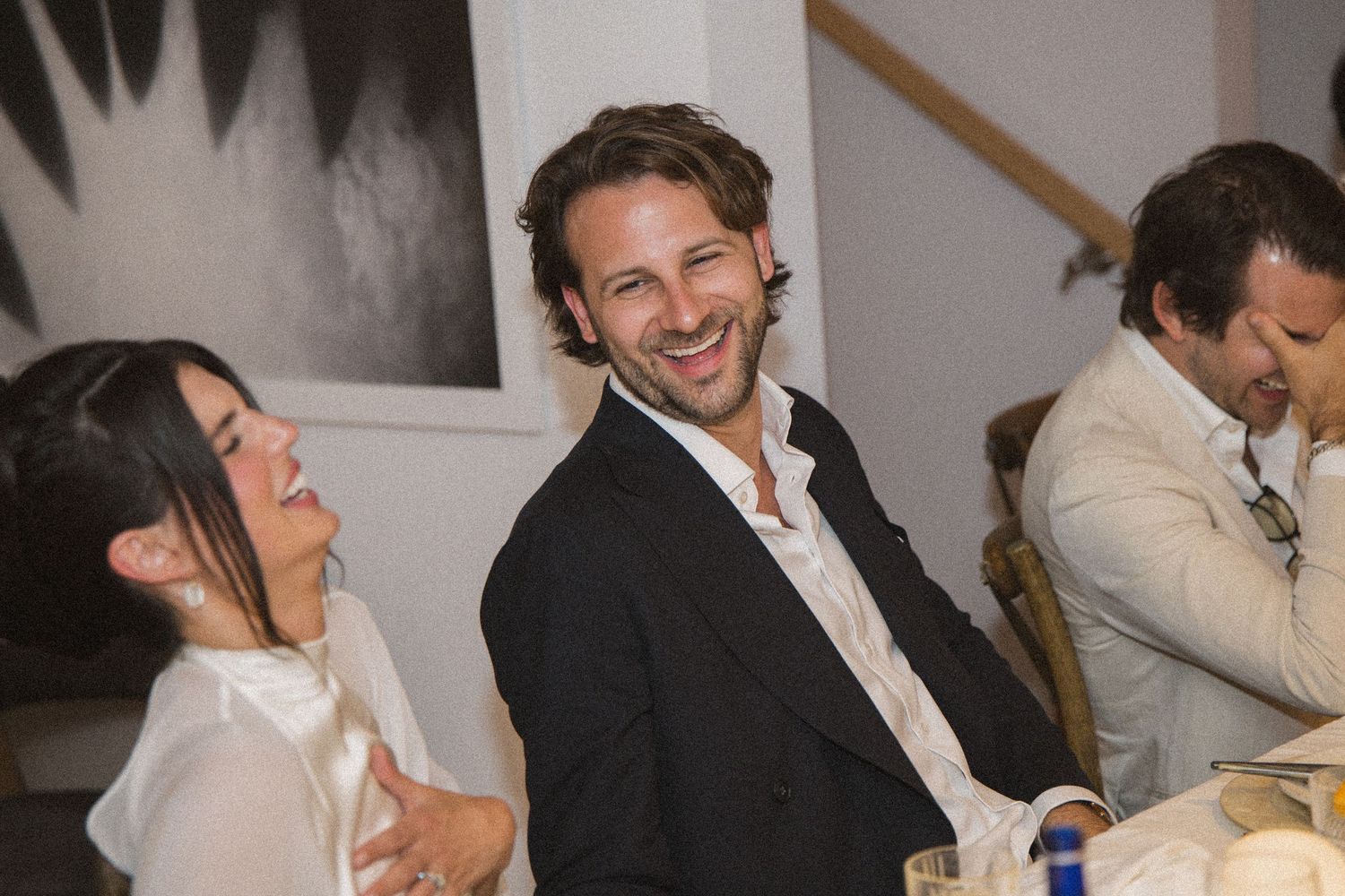 Two guests laughing and smiling at a formal dinner party, man in dark blazer and woman in white outfit.