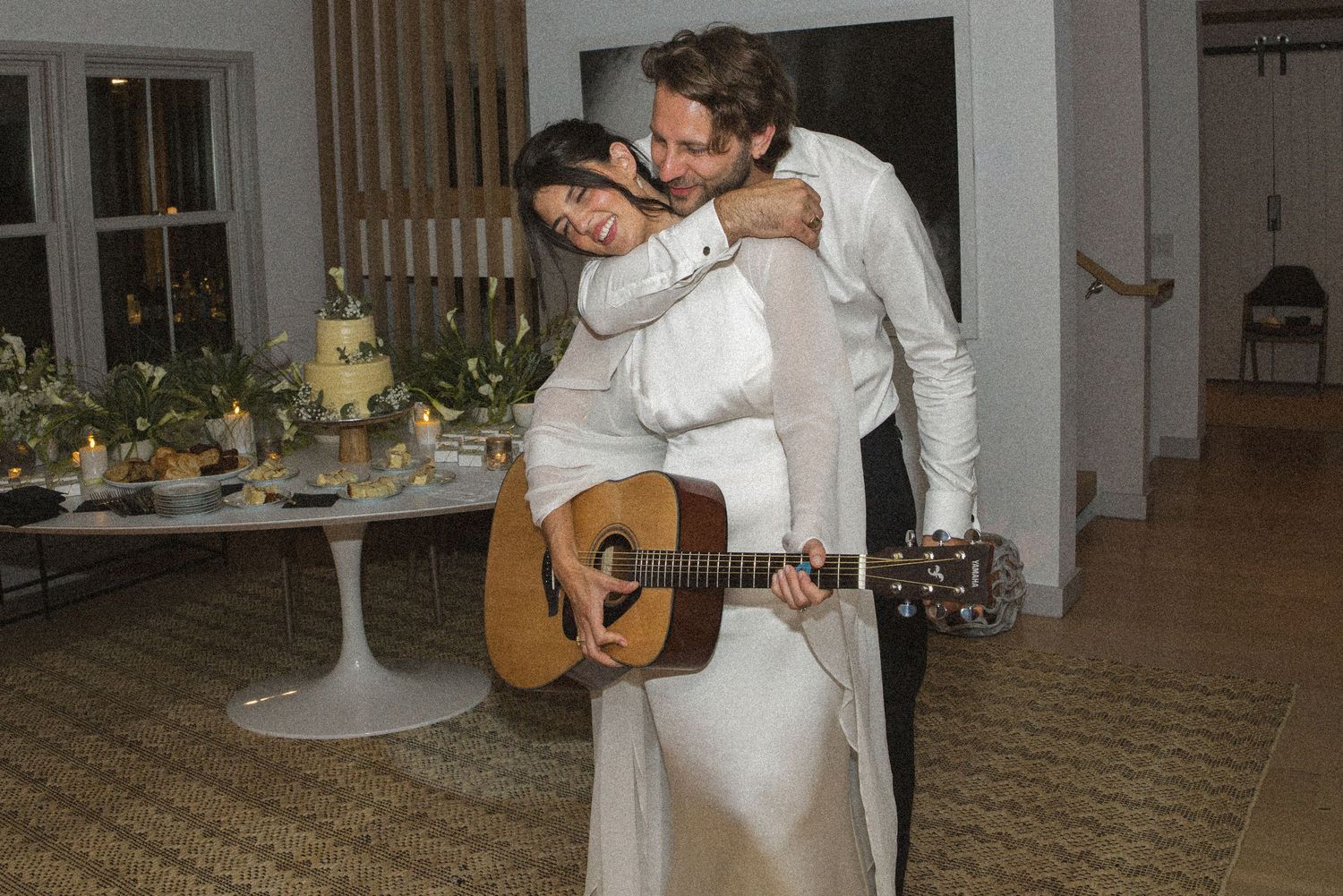 Couple embracing at wedding reception, woman in white gown holding acoustic guitar while man kisses her cheek.