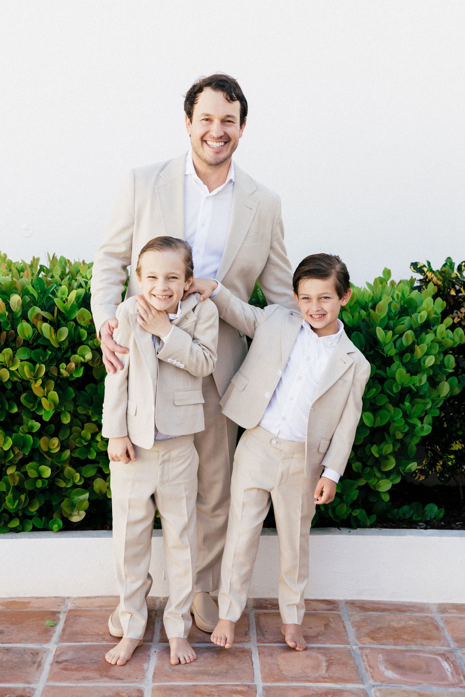 Joyful family portrait with everyone wearing matching beige suits against greenery.