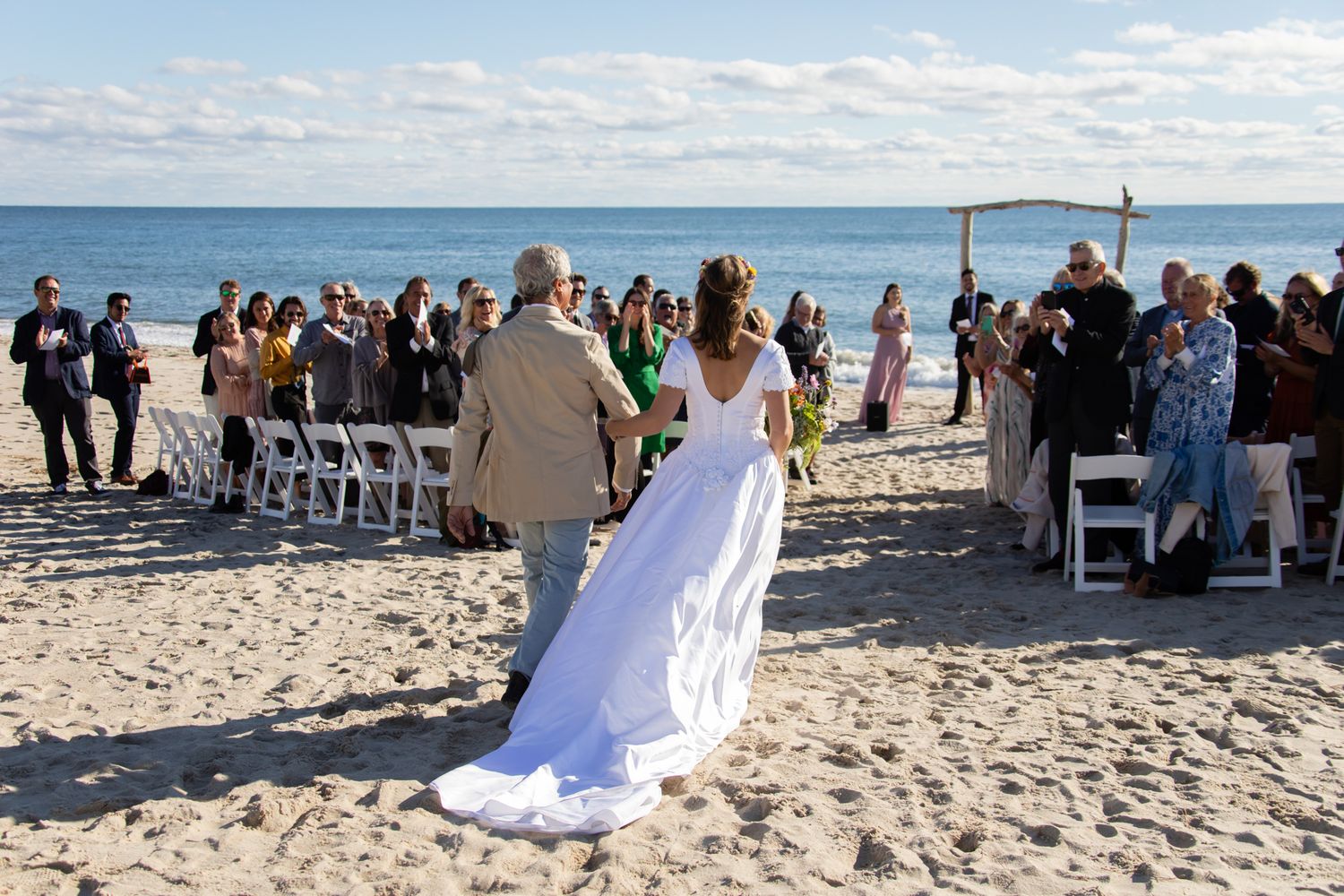 A beach wedding ceremony with guests seated in white chairs along the shoreline.