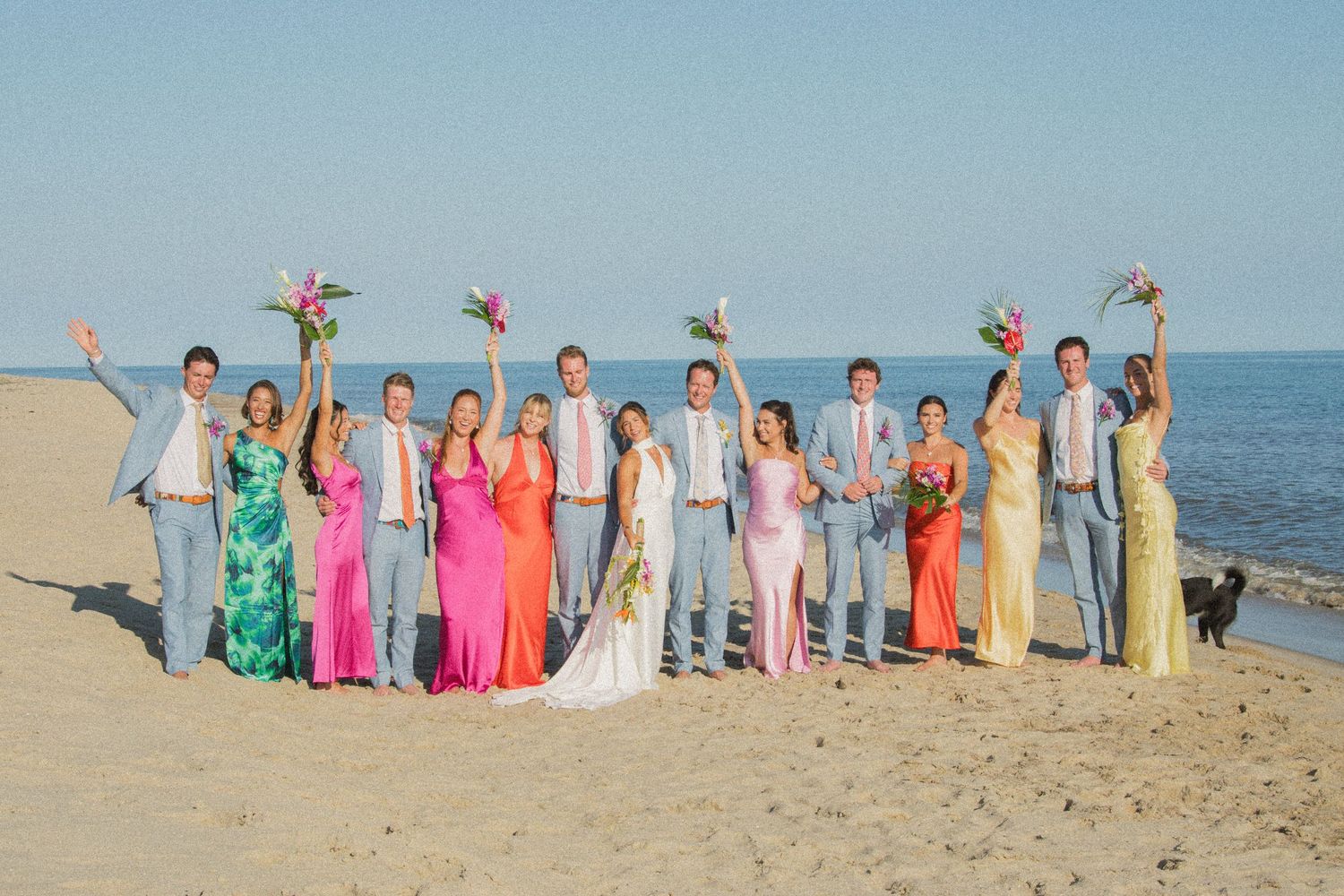 Wedding party in pastel formal attire posing together on a beach with ocean backdrop.
