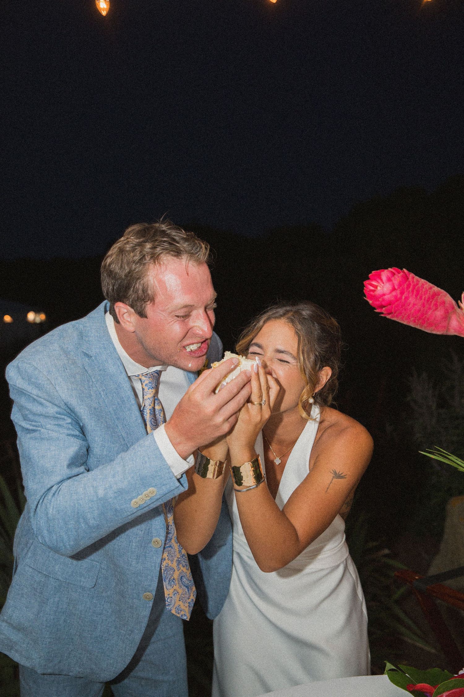 Couple sharing laughter while feeding each other wedding cake at outdoor reception.