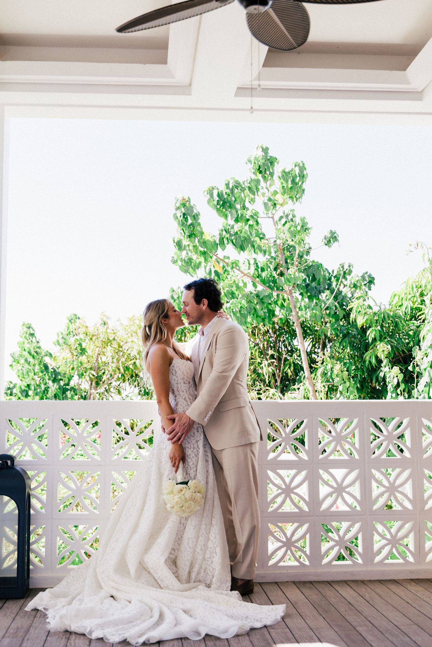A romantic wedding portrait on a white veranda with lush greenery in the background.
