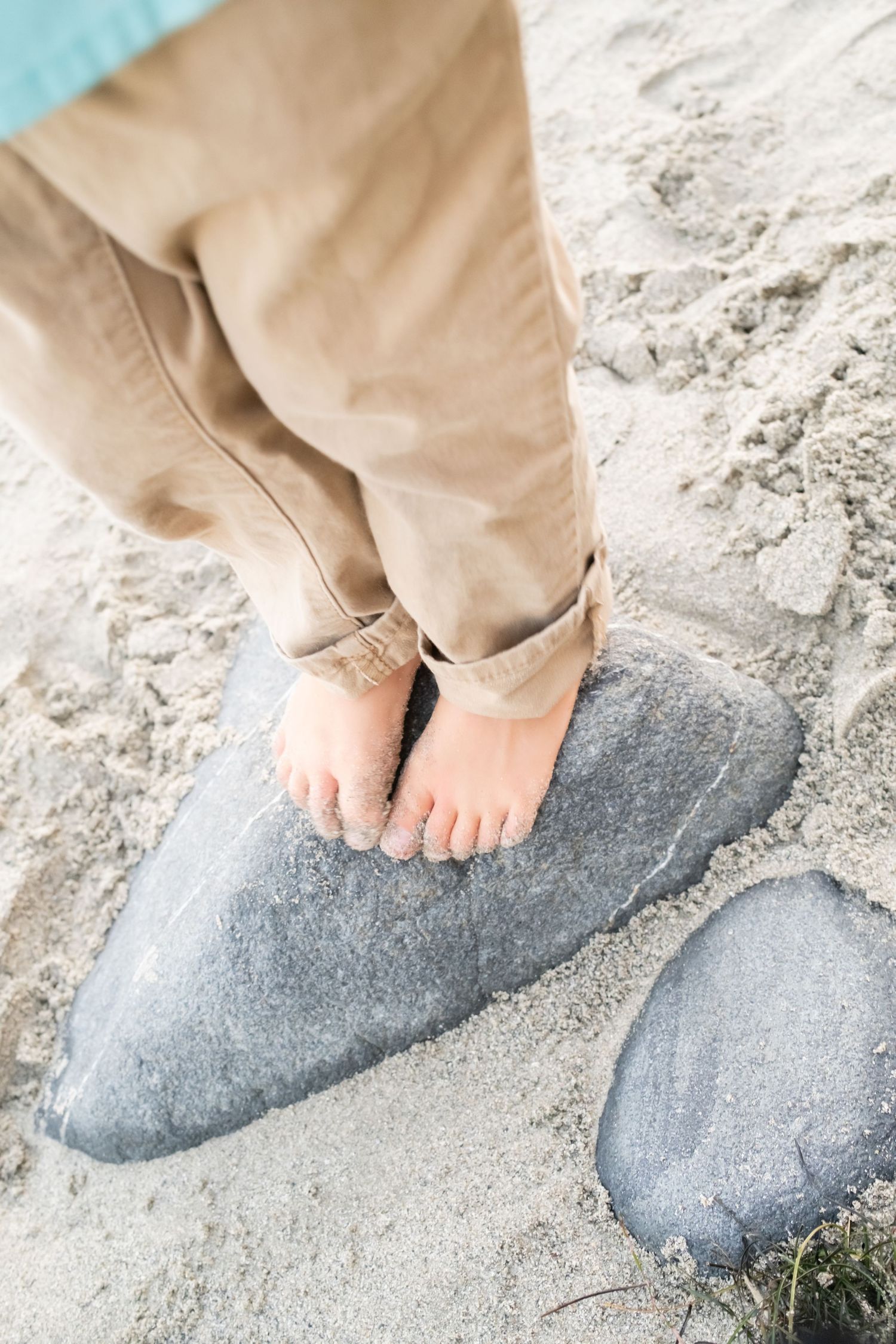 Bare feet in rolled up khaki pants standing on a grey rock with sand.