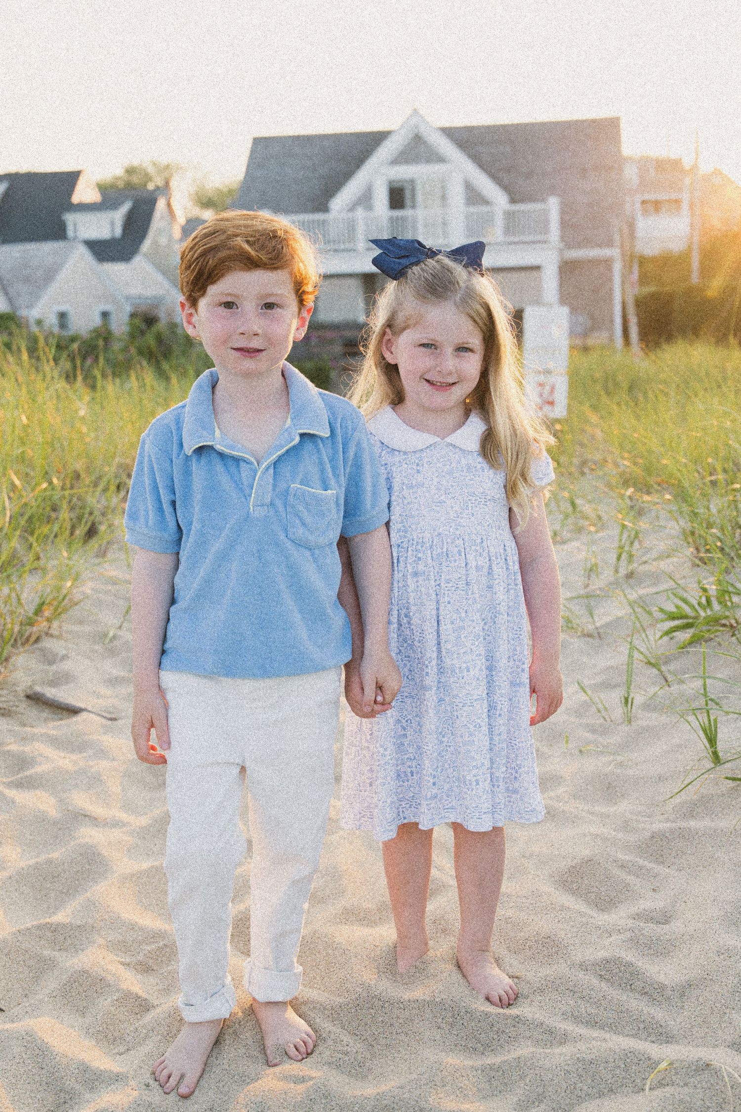 Two young children, a red-haired boy and a girl with a bow, stand barefoot on sandy beach near coastal homes at sunset.