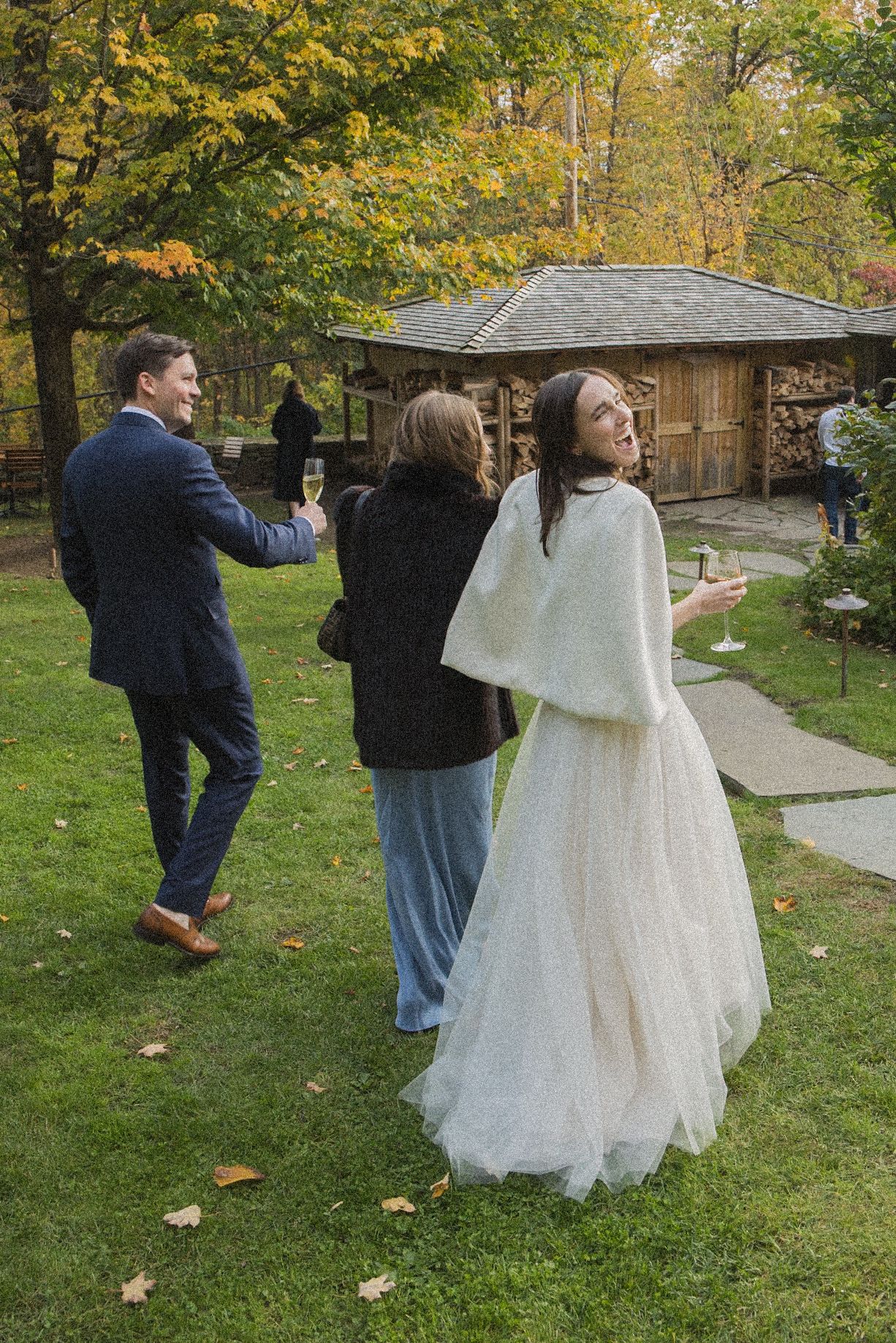 A bride in a white gown and shawl walks across an autumn lawn with a man and woman, all holding champagne glasses near a rustic cabin.