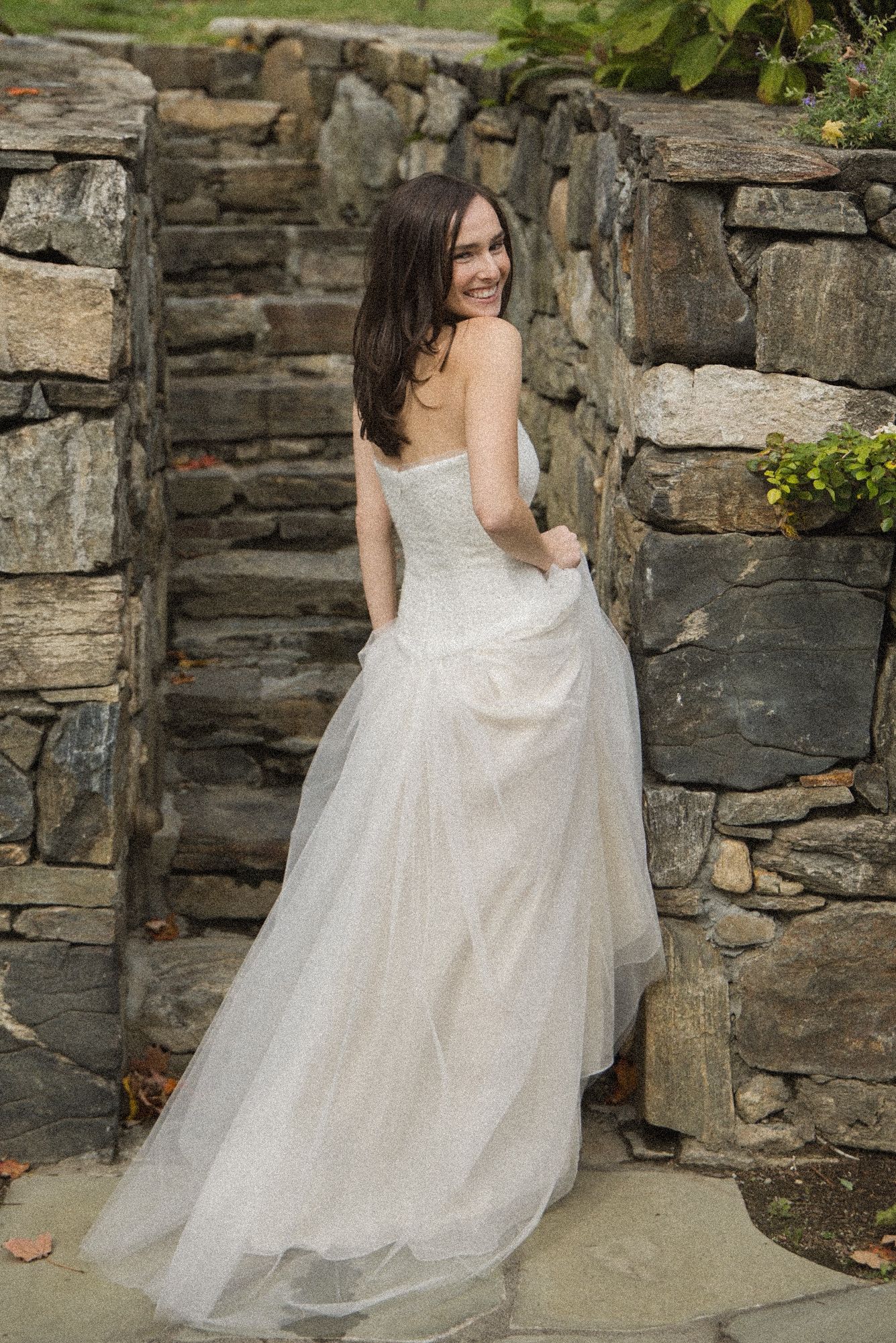 Smiling bride in a strapless white tulle wedding gown looks over her shoulder beside a rustic stone wall and steps.
