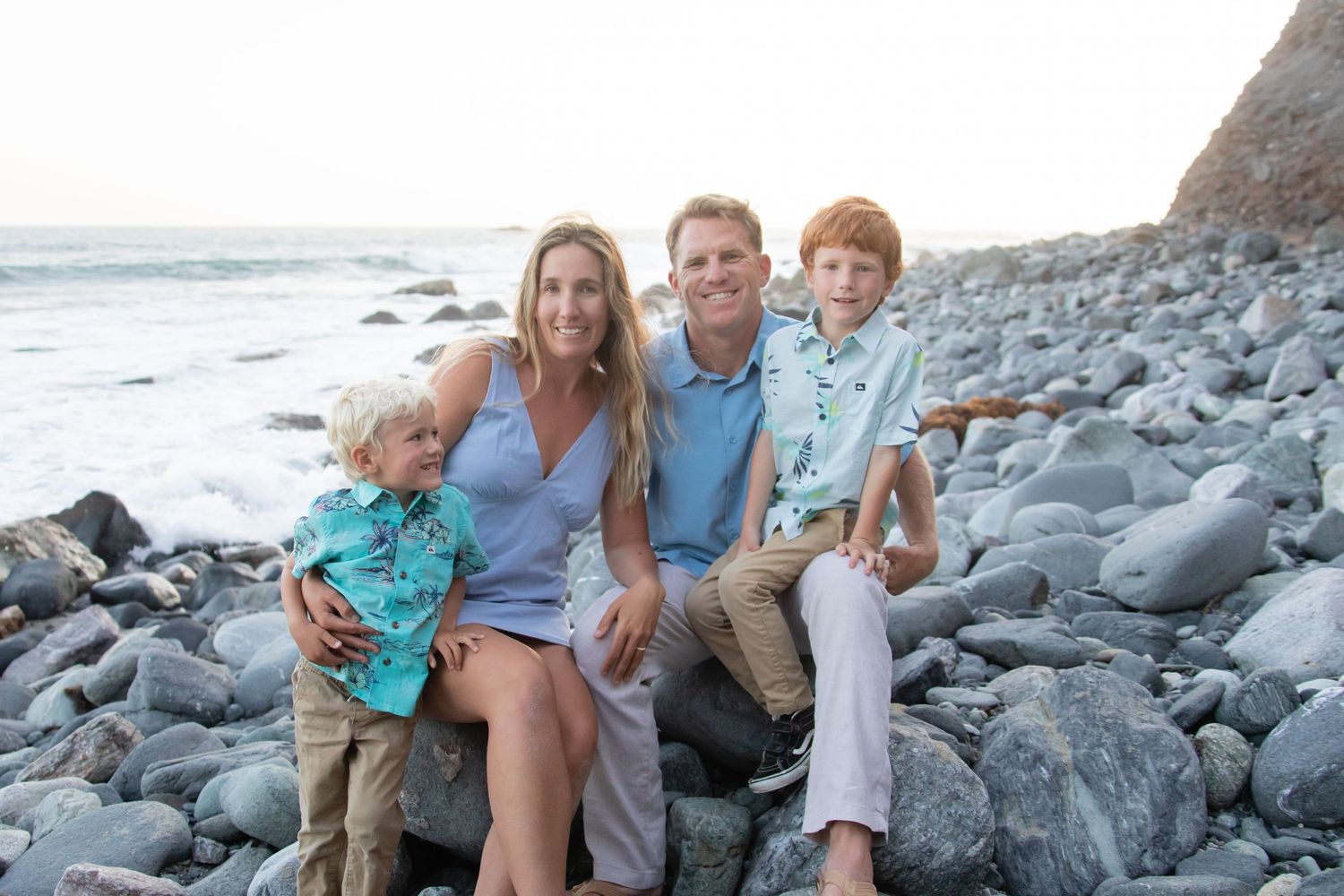 A family sits together on rocky coastal beach during sunset with ocean waves in background.