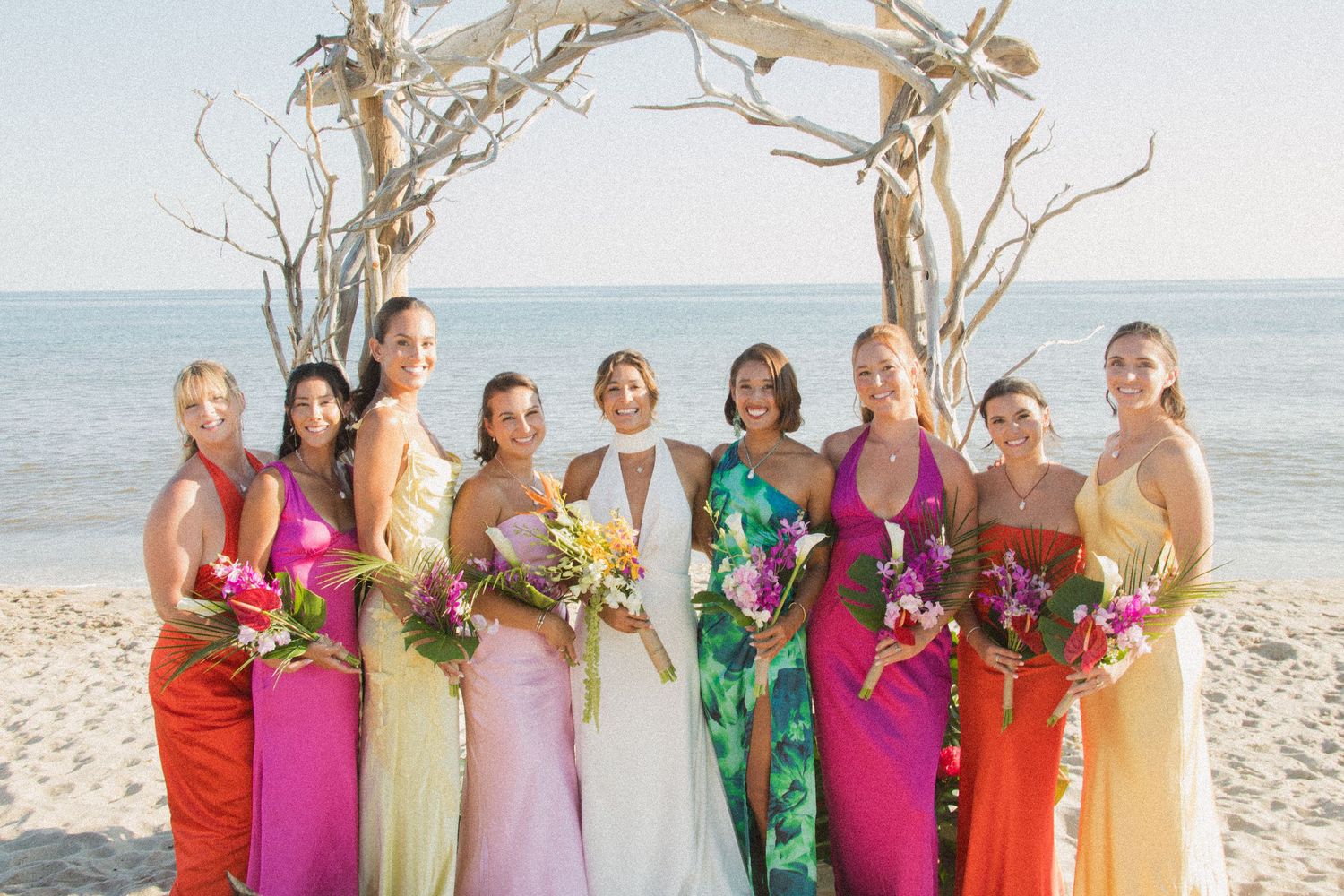 Bridesmaids in vibrant rainbow-colored dresses pose with bride under beach arch.