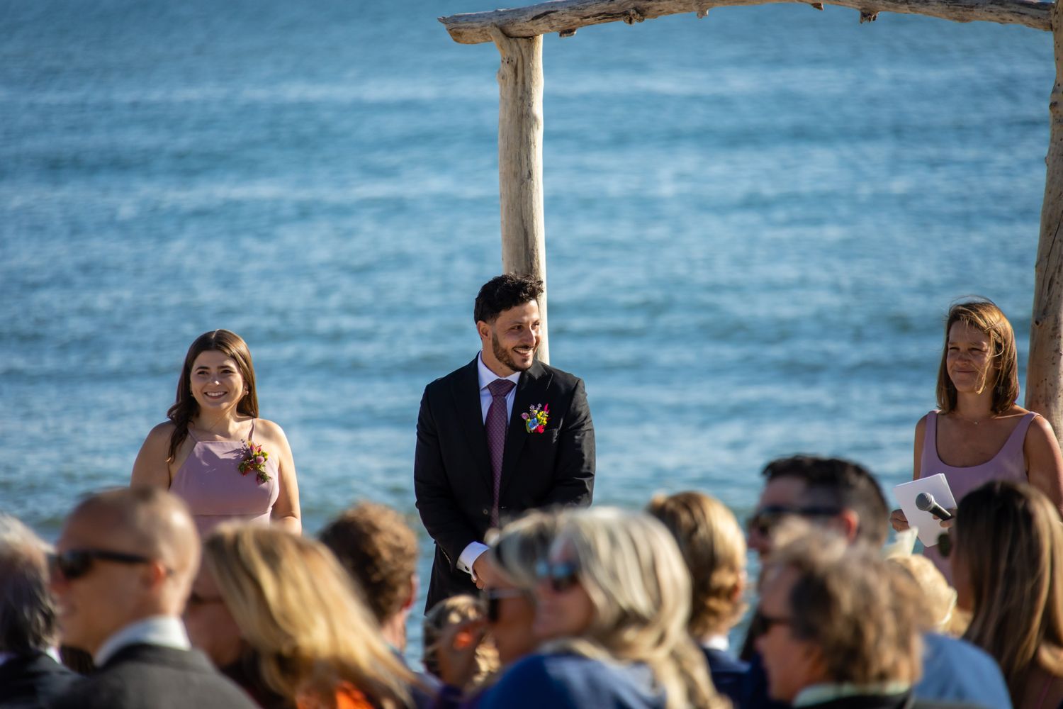 Beach wedding ceremony under driftwood arch with guests seated and ocean views.