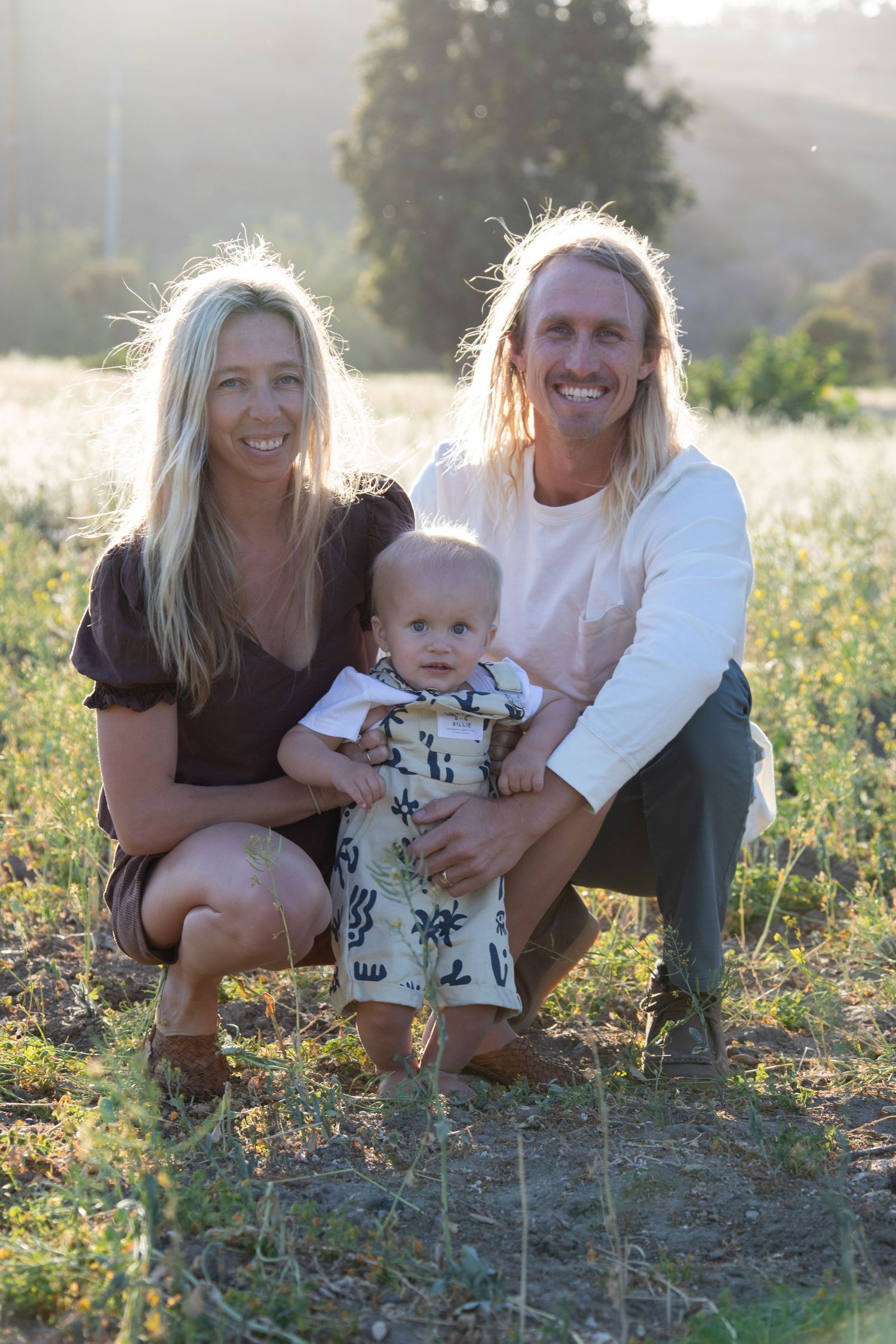A family of three sits together in a golden sunset meadow.