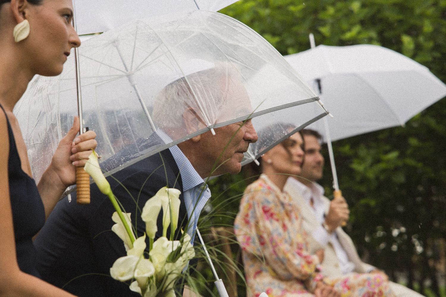 Guests holding clear umbrellas at an outdoor wedding ceremony on a rainy day, with white calla lily bouquet.