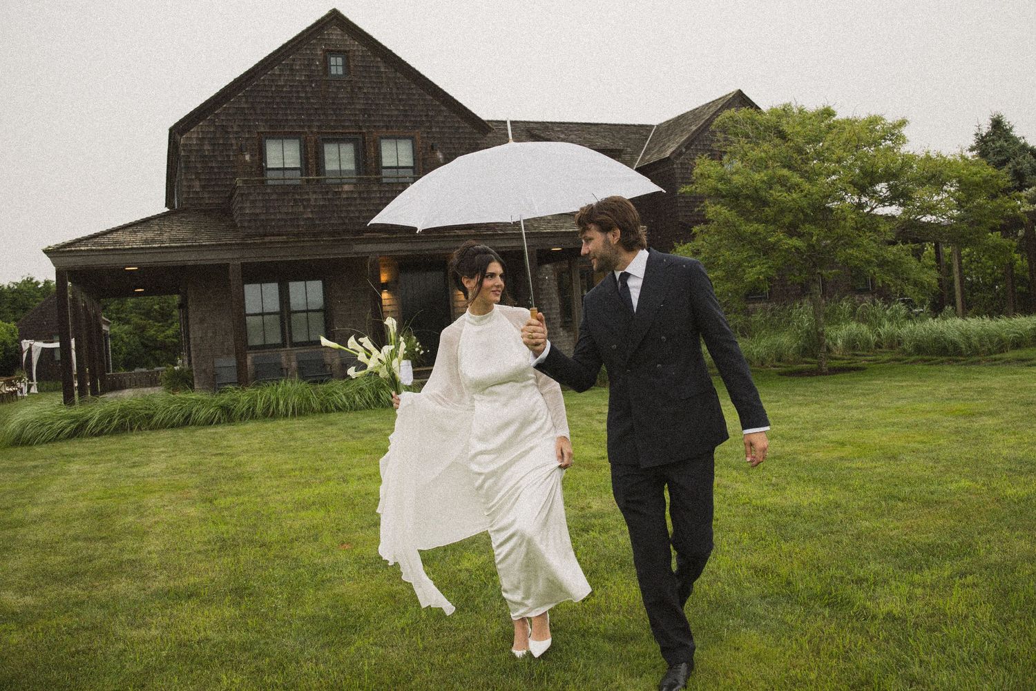 Bride and groom walking under white umbrella on lush green lawn outside rustic brown house on rainy wedding day.