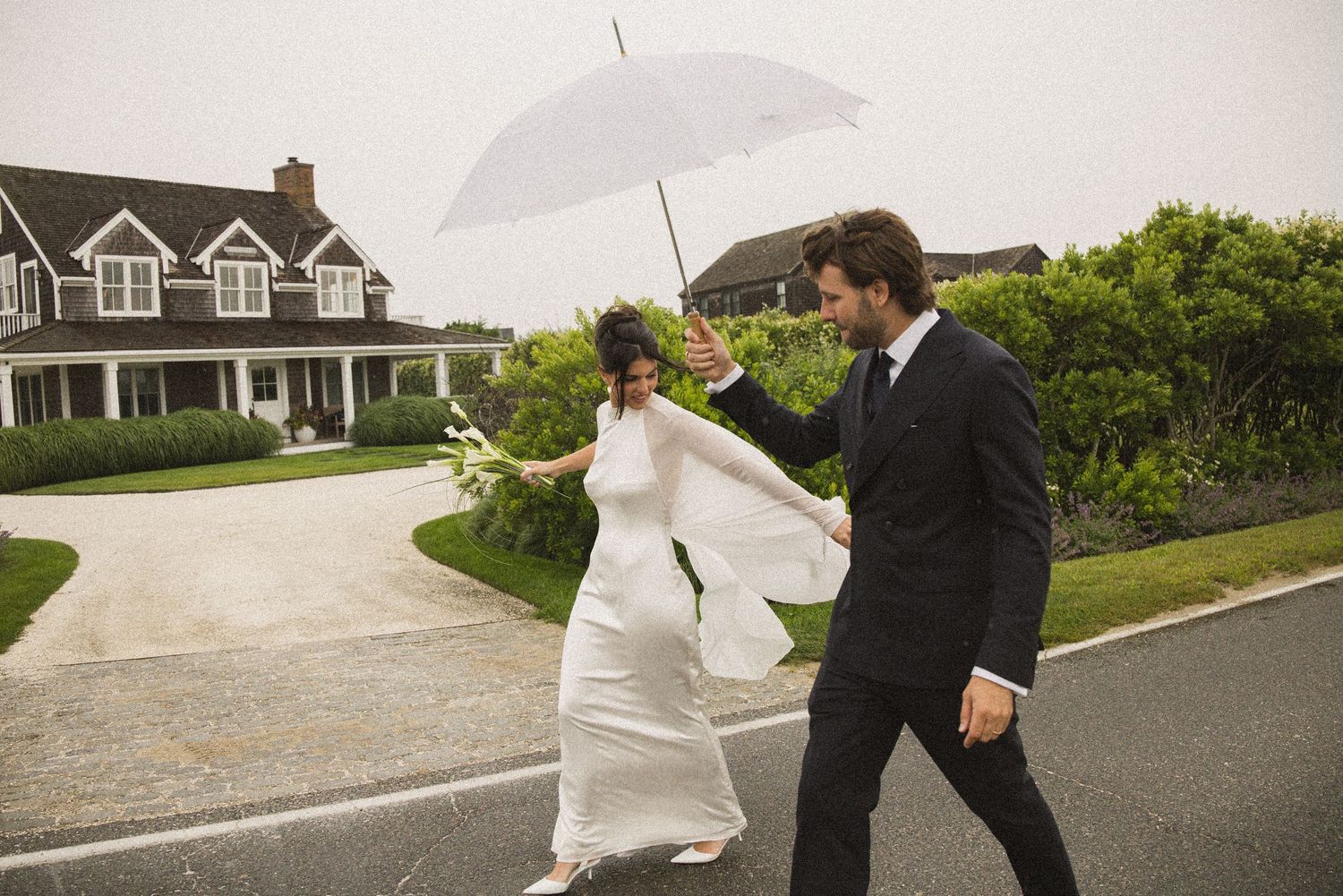 Bride and groom walking in the rain under a white umbrella on their wedding day outside a elegant home.