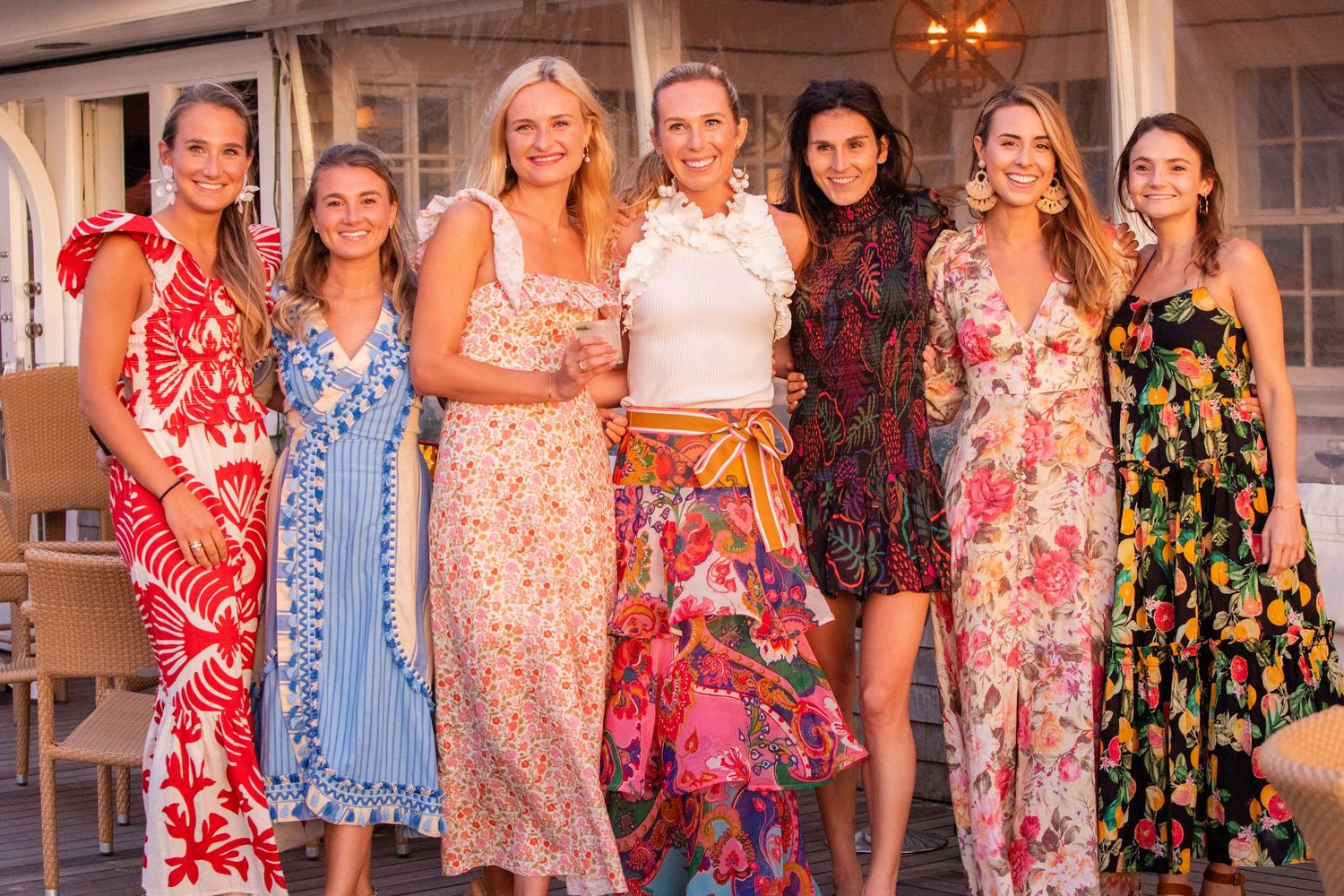 Seven women in colorful floral dresses smiling together at an outdoor summer gathering.