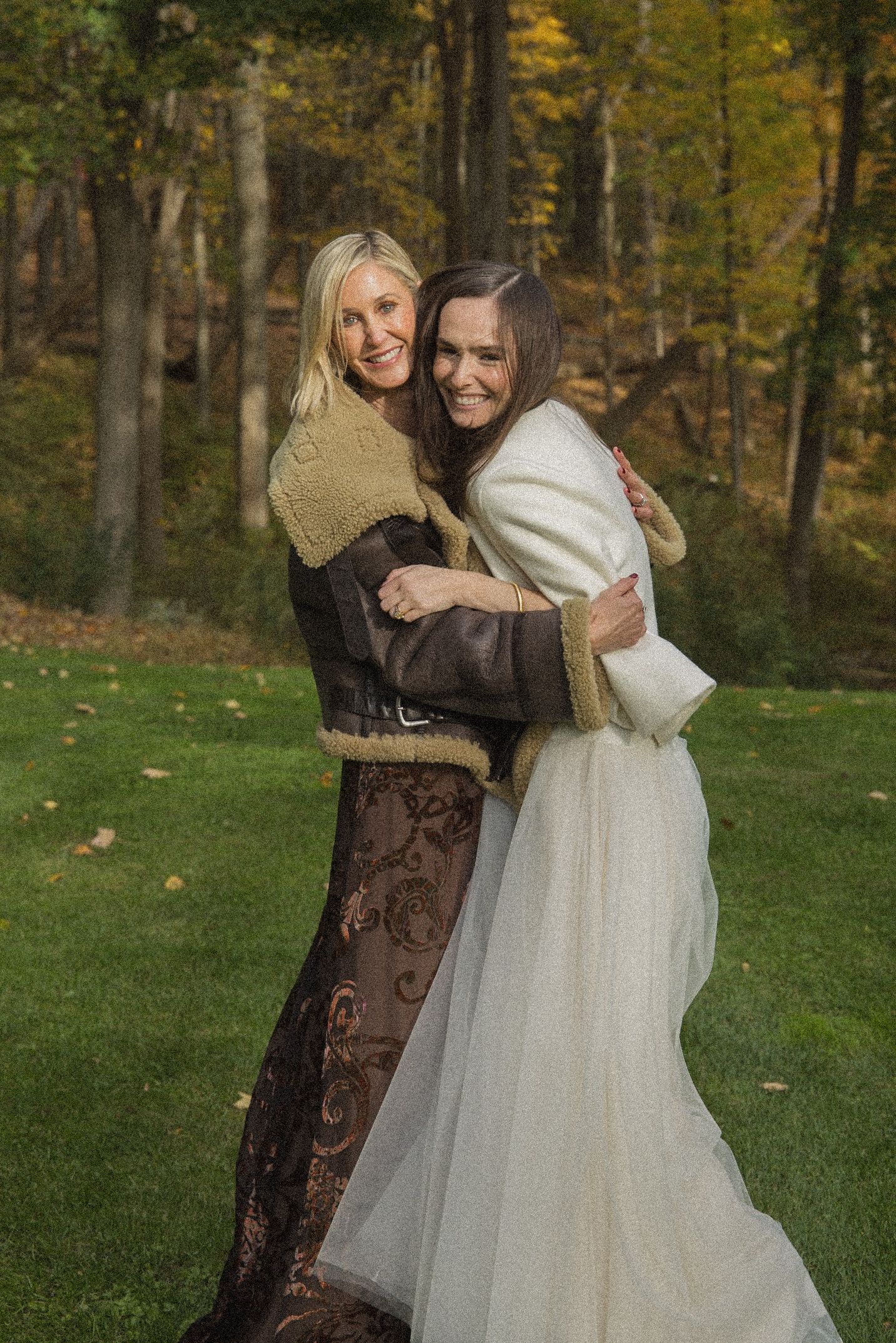 Two smiling women embrace outdoors on a green lawn, one in a shearling jacket and brown dress, the other in a white bridal gown.