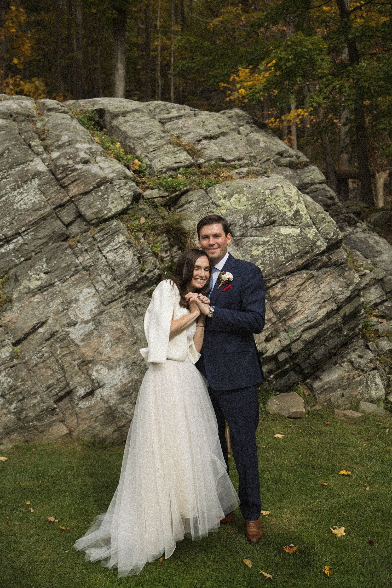 Smiling bride in a white tulle skirt and groom in a navy suit embrace in front of a large mossy boulder on an autumn lawn.