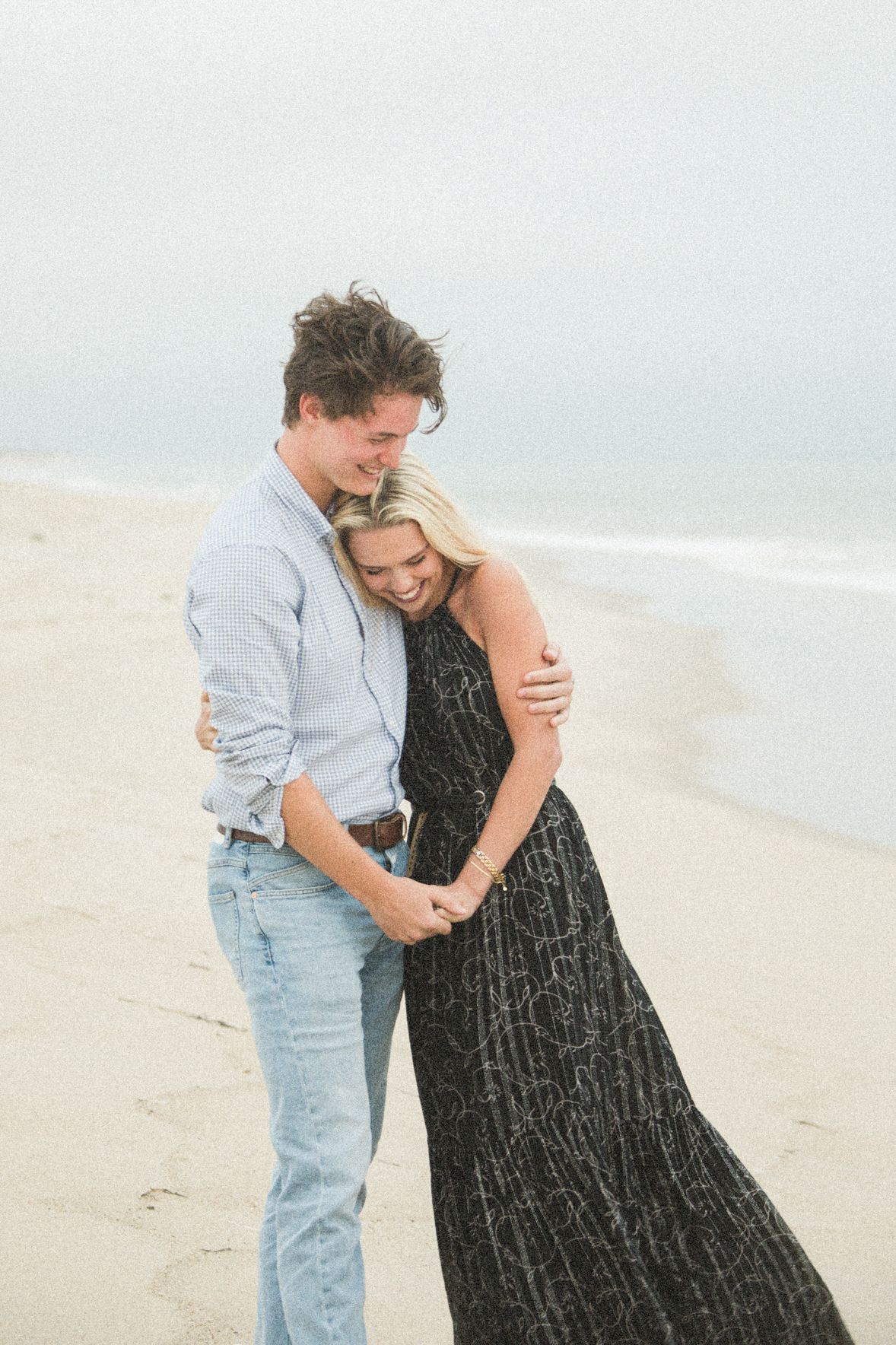 A couple embraces and laughs on a sandy beach, woman in black maxi dress and man in light blue shirt and jeans.